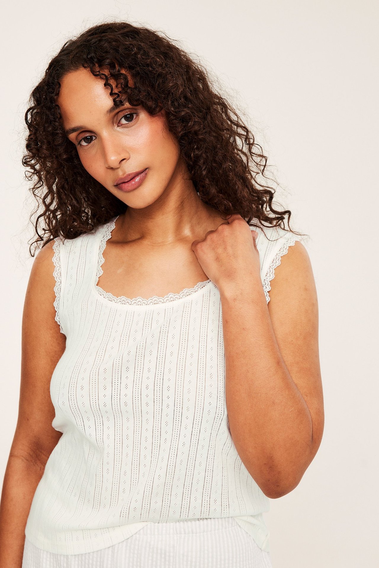 Nærbilde av kvinne i offwhite pointelle pyjamas topp med blondekant, stylet med lyse stripete underdeler.