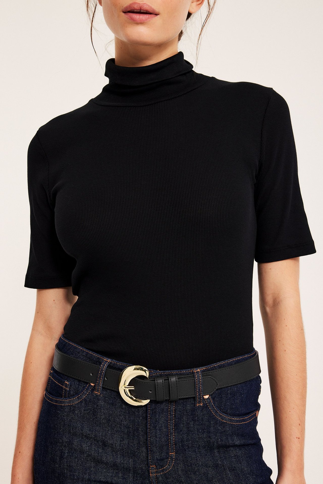 Närbild, framifrån: Dam i svart bälte av fuskläder med stort guldfärgat spänne, mörkblå jeans och svart ribbad polotröja.