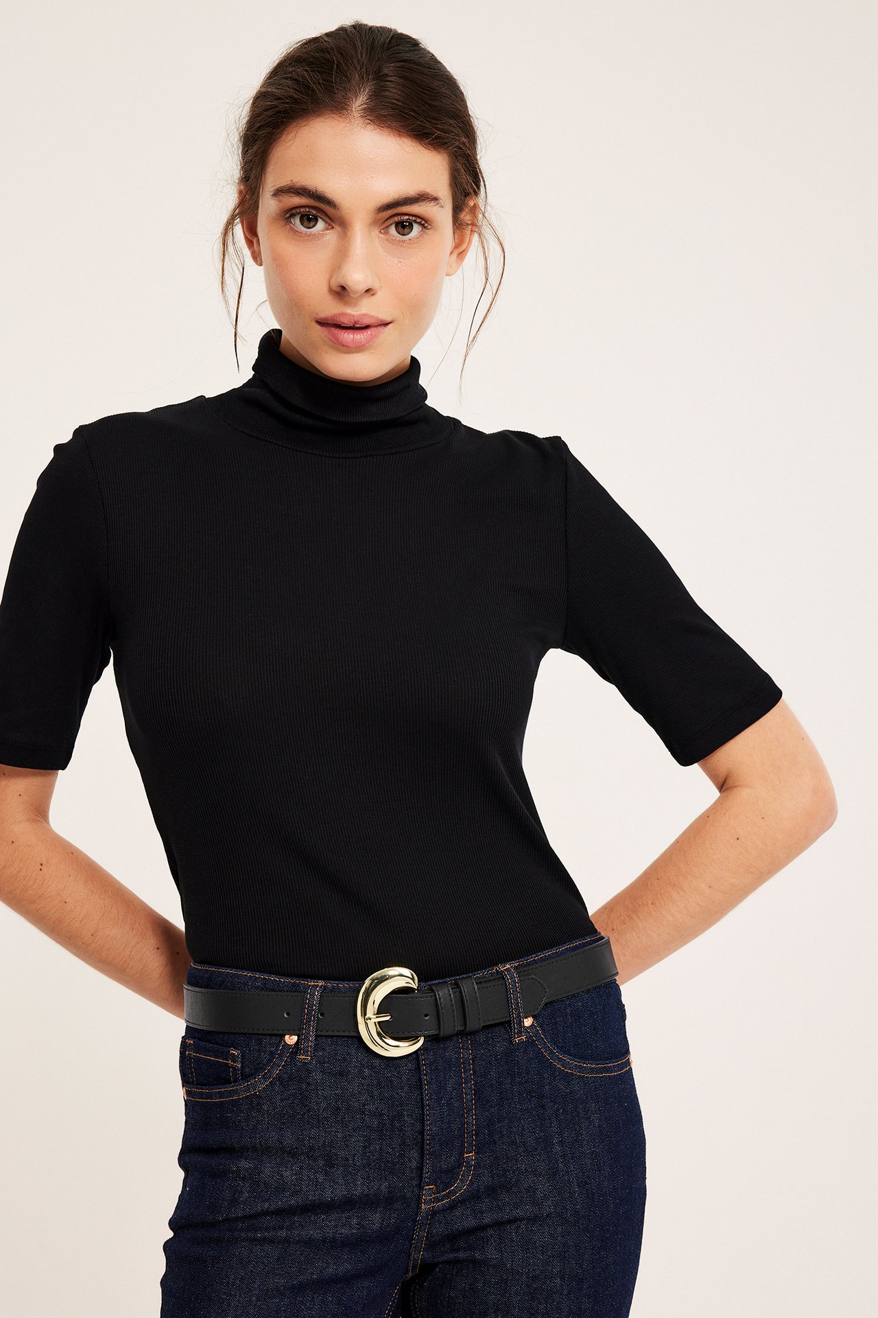 Front view: Kvinna med svart fuskläderbälte, guldspänne, svart ribbad polotröja, mörka jeans.