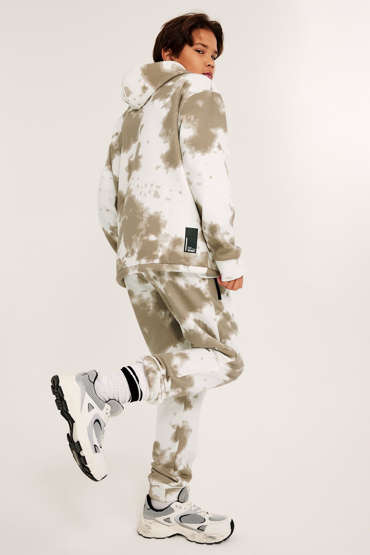 Poikapotilaan takaa näkymä valko-beigessä solmuvärjätyssä hupparissa, yhteensopivissa collegehousuissa, valkoisissa sukissa ja lenkkareissa.