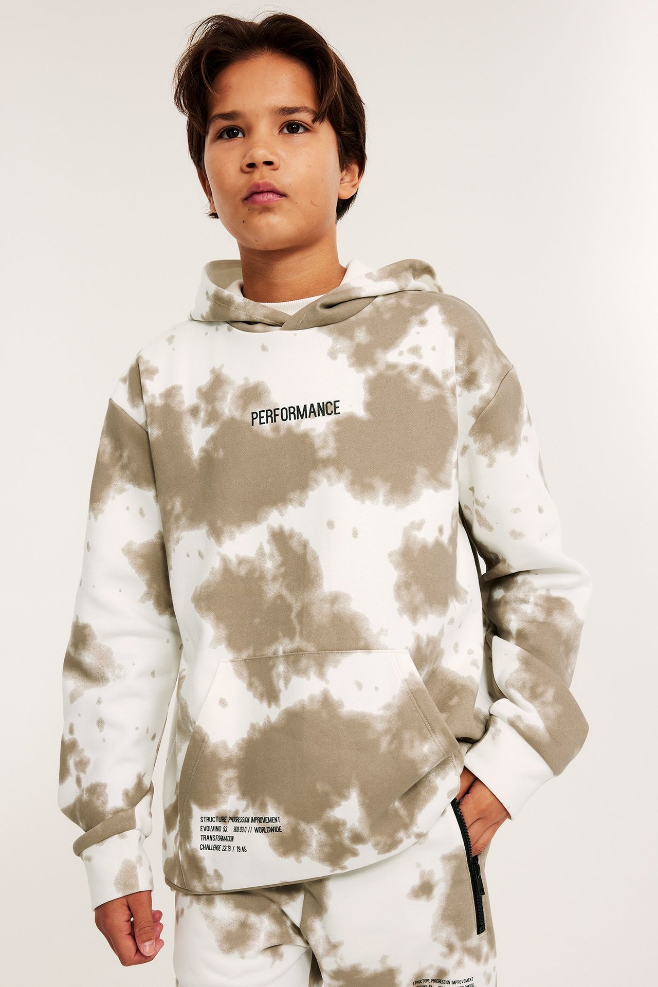 Edestäpäin kuvattuna lapsi valko-beigessä solmuvärjätyssä hupparissa, jossa on kengurutasku ja yhteensopivat collegehousut.