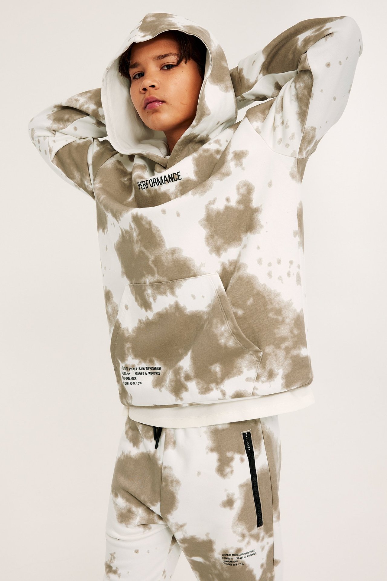 Edestä näkymä lapsesta valko-beigessä solmuvärjätyssä hupparissa kängurutaskulla ja yhteensopivissa collegehousuissa.