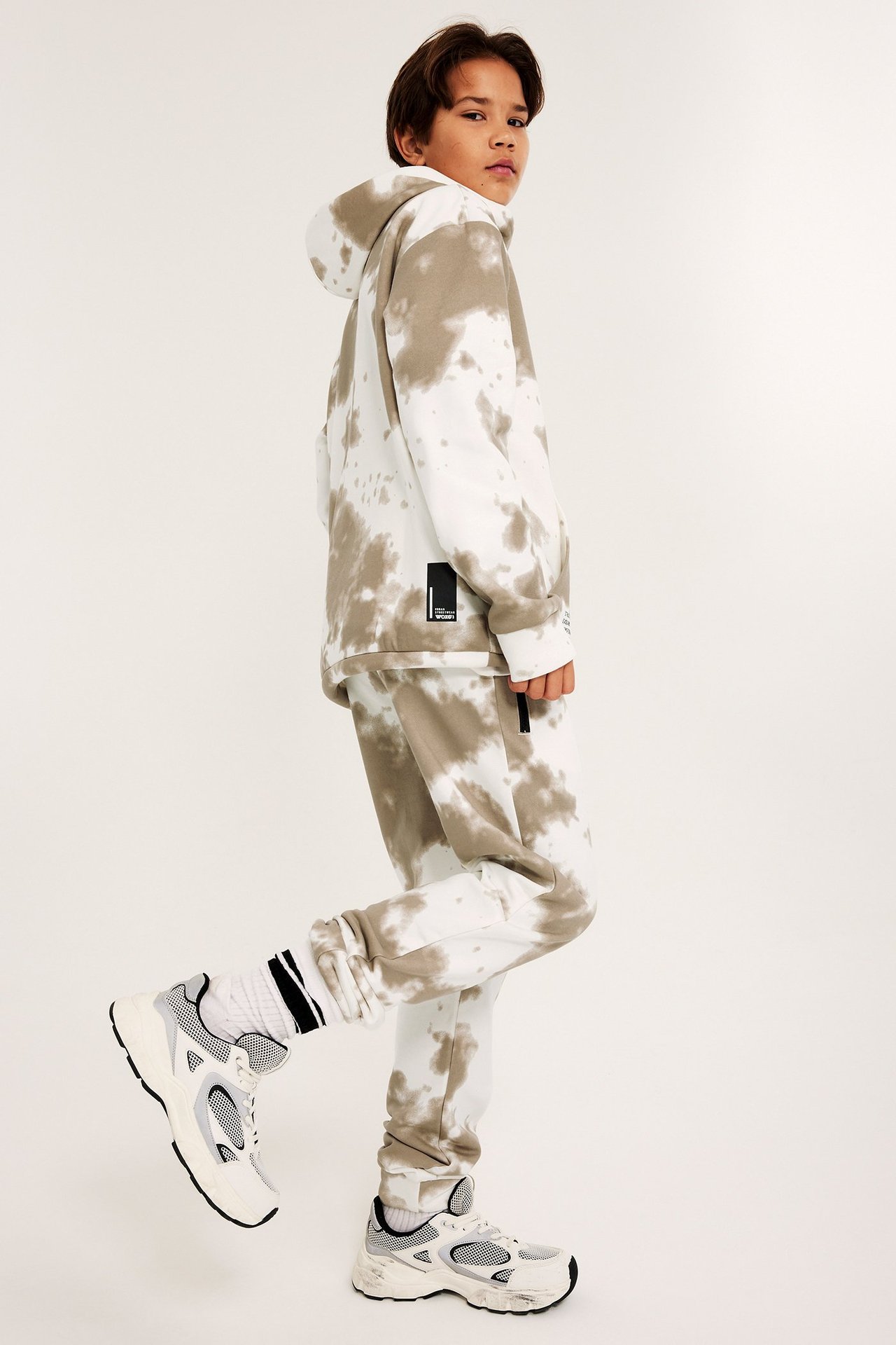 Lapsi sivukuvassa valko-beigeissä kuviollisissa verryttelyhousuissa, hupparissa, sukissa ja lenkkareissa.