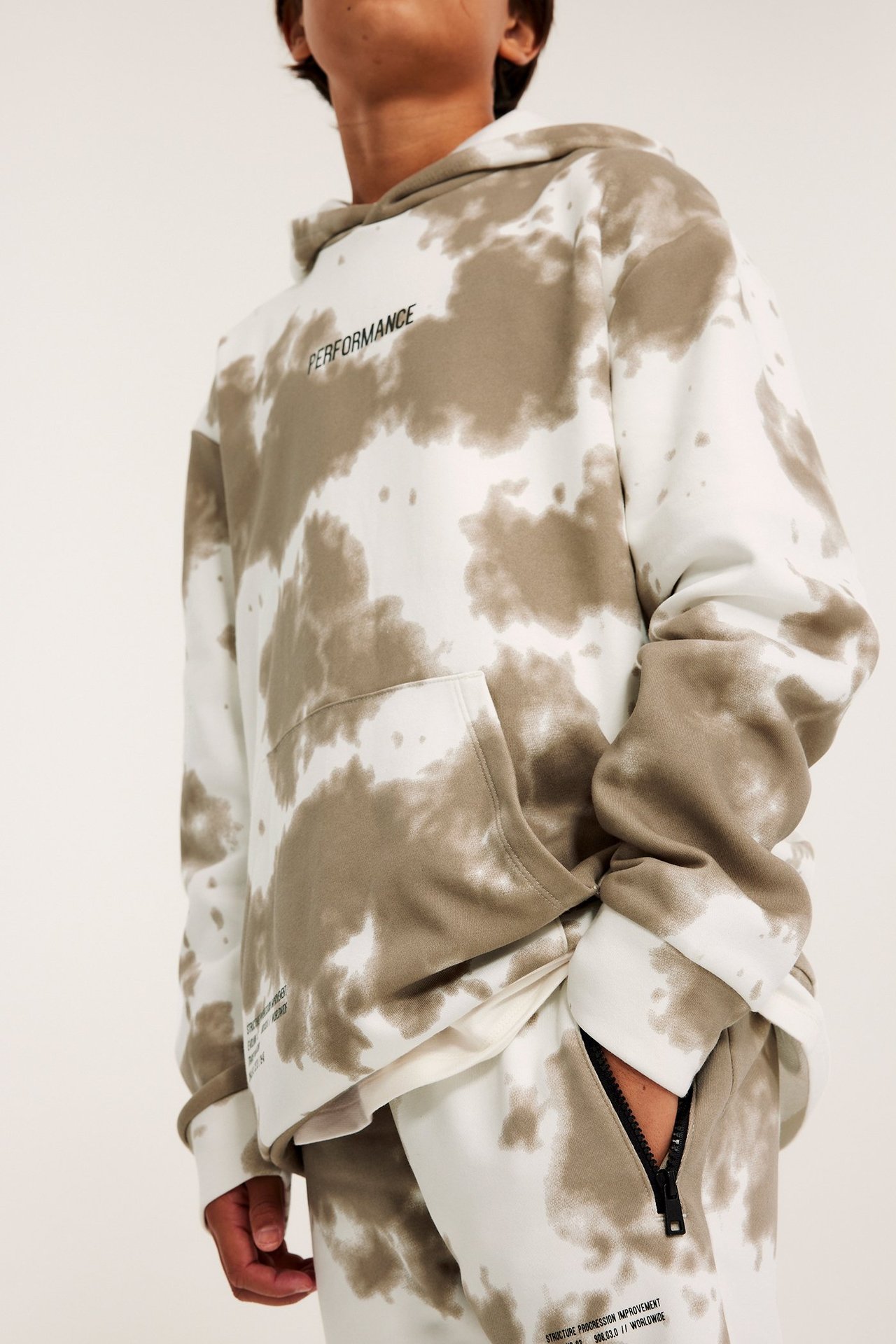 Edestäpäin kuvassa lapsi valkoisissa ja ruskeissa tie-dye collegehousuissa vetoketjutaskulla ja yhteensopivassa hupparissa.