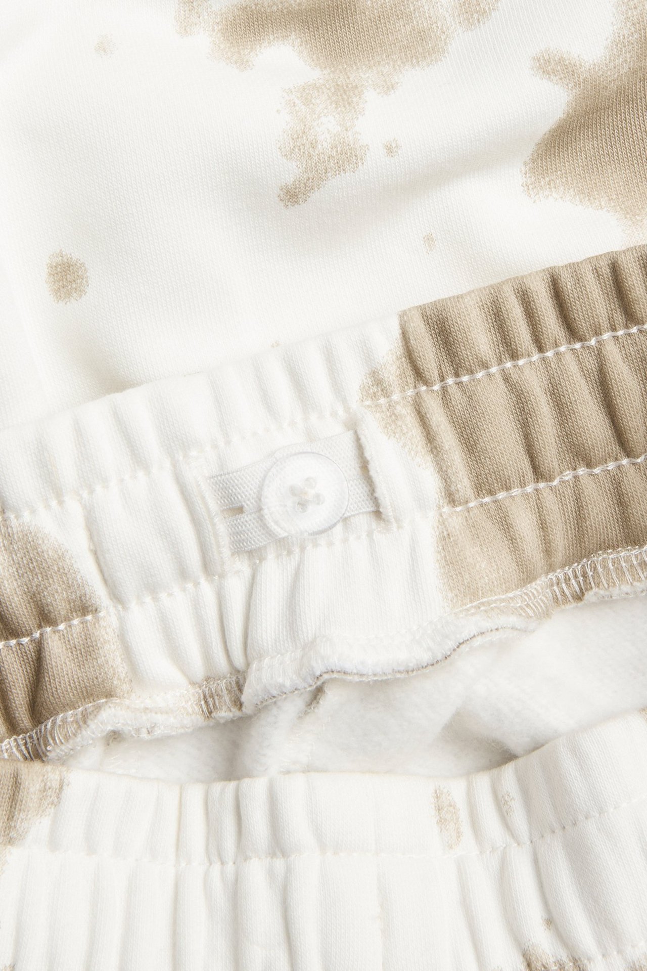 Lähikuva valko-beigekuvioisista verryttelyhousuista, joissa joustava vyötärö ja nappisäätö.