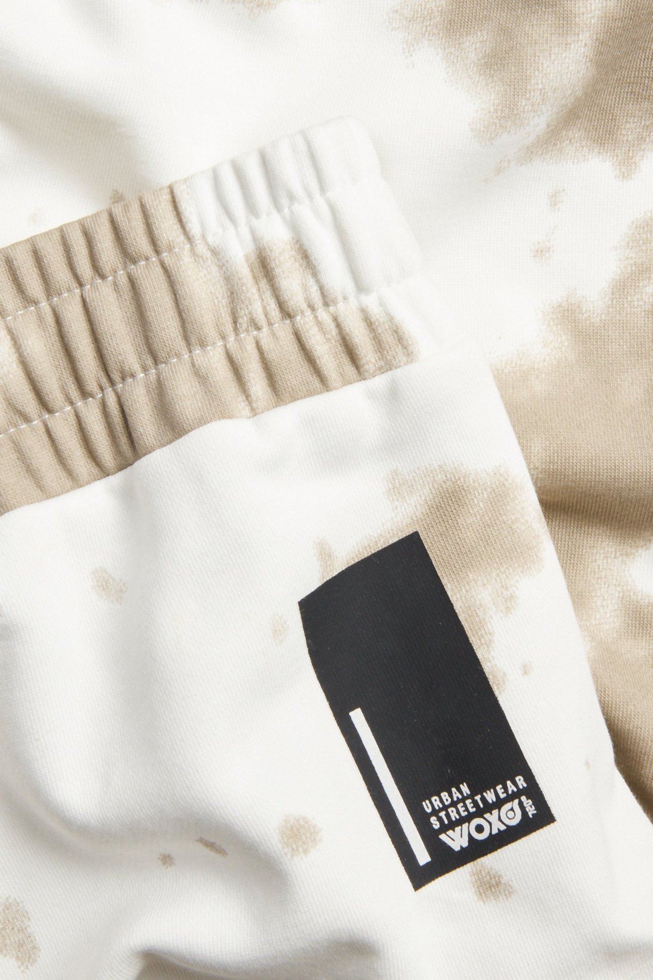 Lähikuva valkoisista ja beigenvärisistä kuviollisista lasten collegehousuista, joissa joustovyötärö ja musta Woxo-etiketti.
