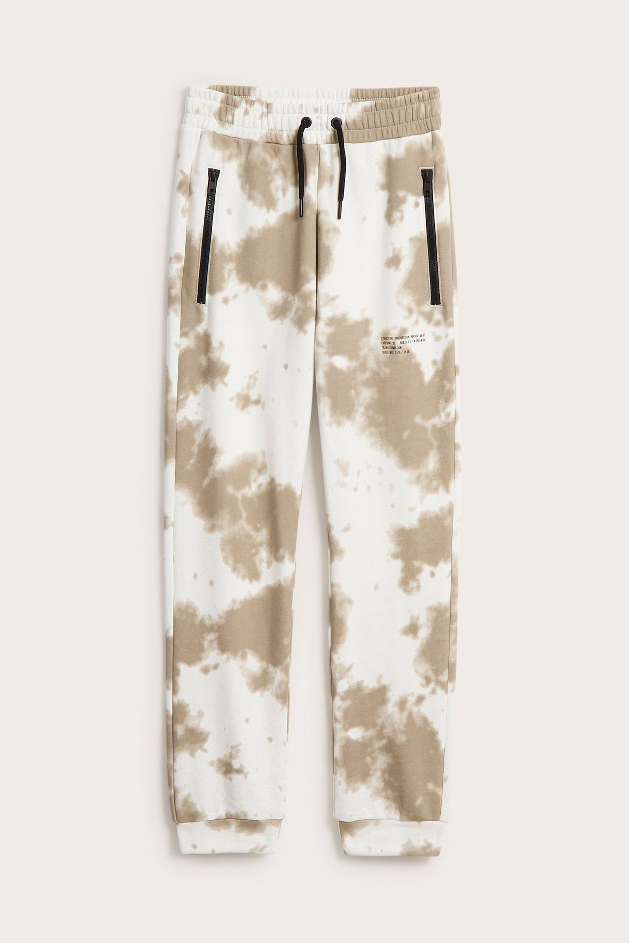 Edestä kuvatut lasten kuviolliset collegehousut, valkoinen ja beige batikkuvio, mustat kiristysnyörit, vetoketjutaskut.