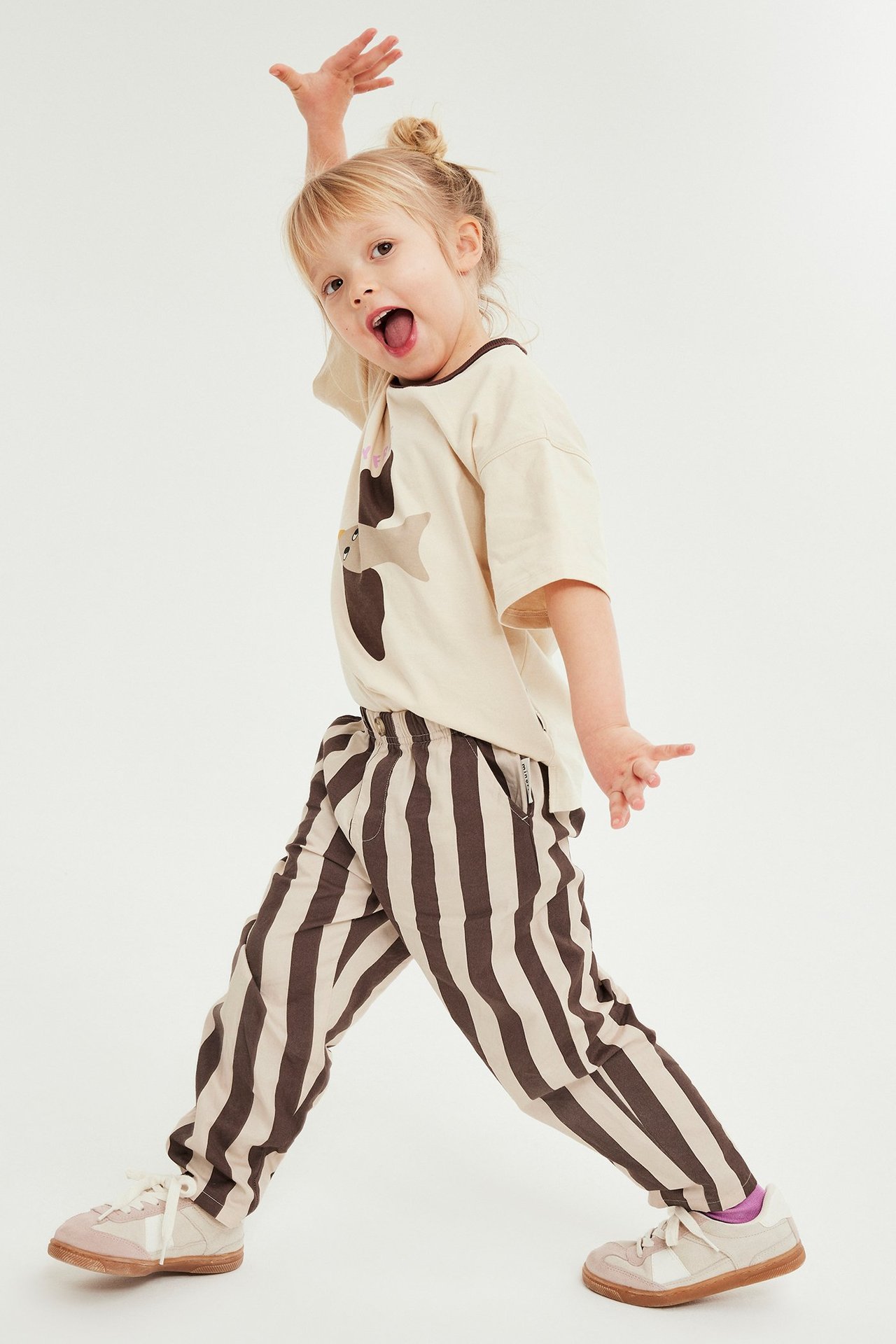 Forfra: barn i brun- og beigestripete vevde joggebukser med lommer, en beige t-skjorte og joggesko.