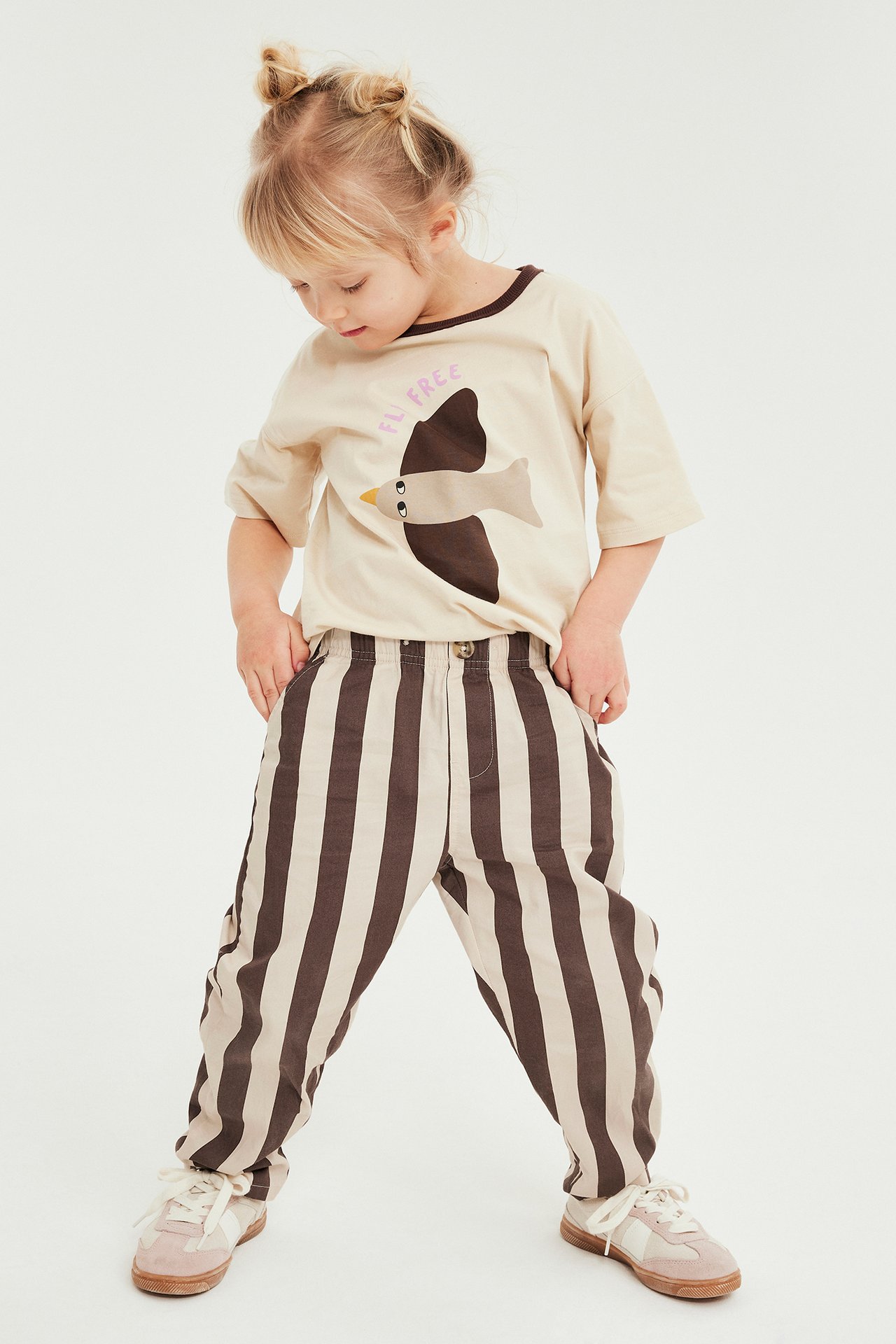 Forfra: barn i brunt og beigefarget stripete bomullsbukse og en beige t-skjorte med fugletrykk.