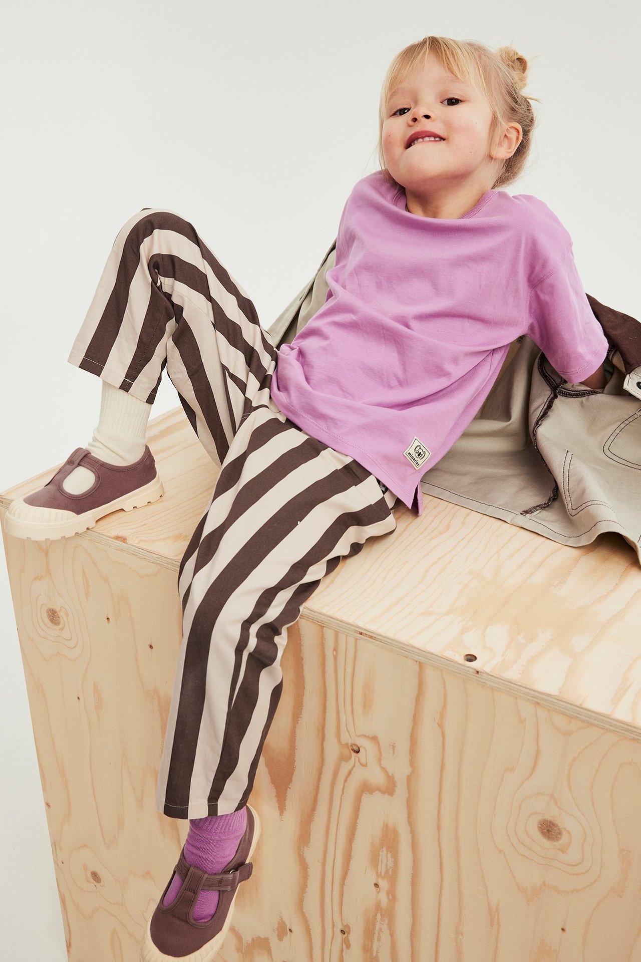 Edestäpäin lapsi ruskeissa raidallisissa puuvillahousuissa, violetissa t-paidassa, ruskeissa kengissä ja sukissa.