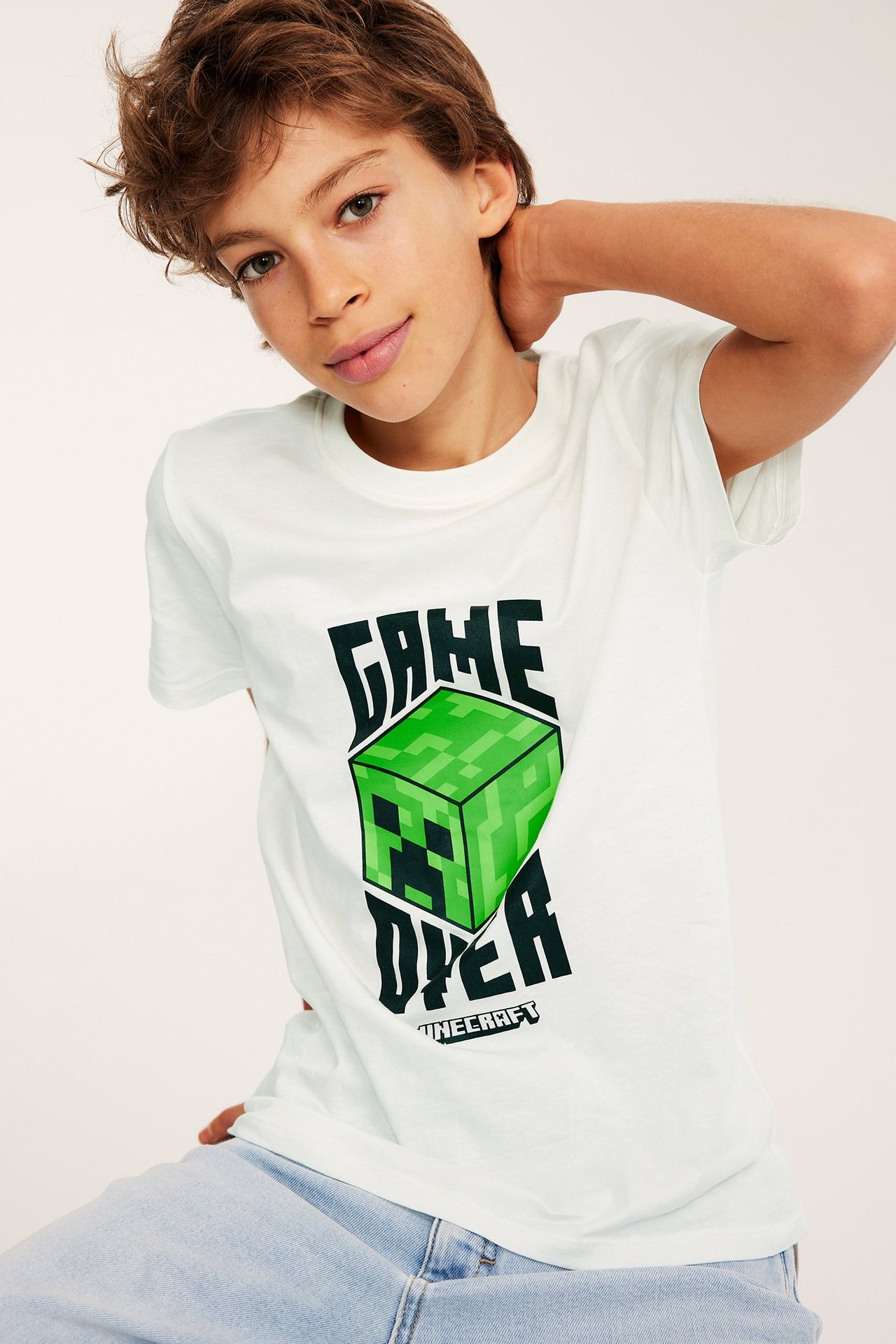 Edestä: poika luonnonvalkoisessa Minecraft-painatetussa puuvilla T-paidassa ja vaaleansinisissä farkuissa.