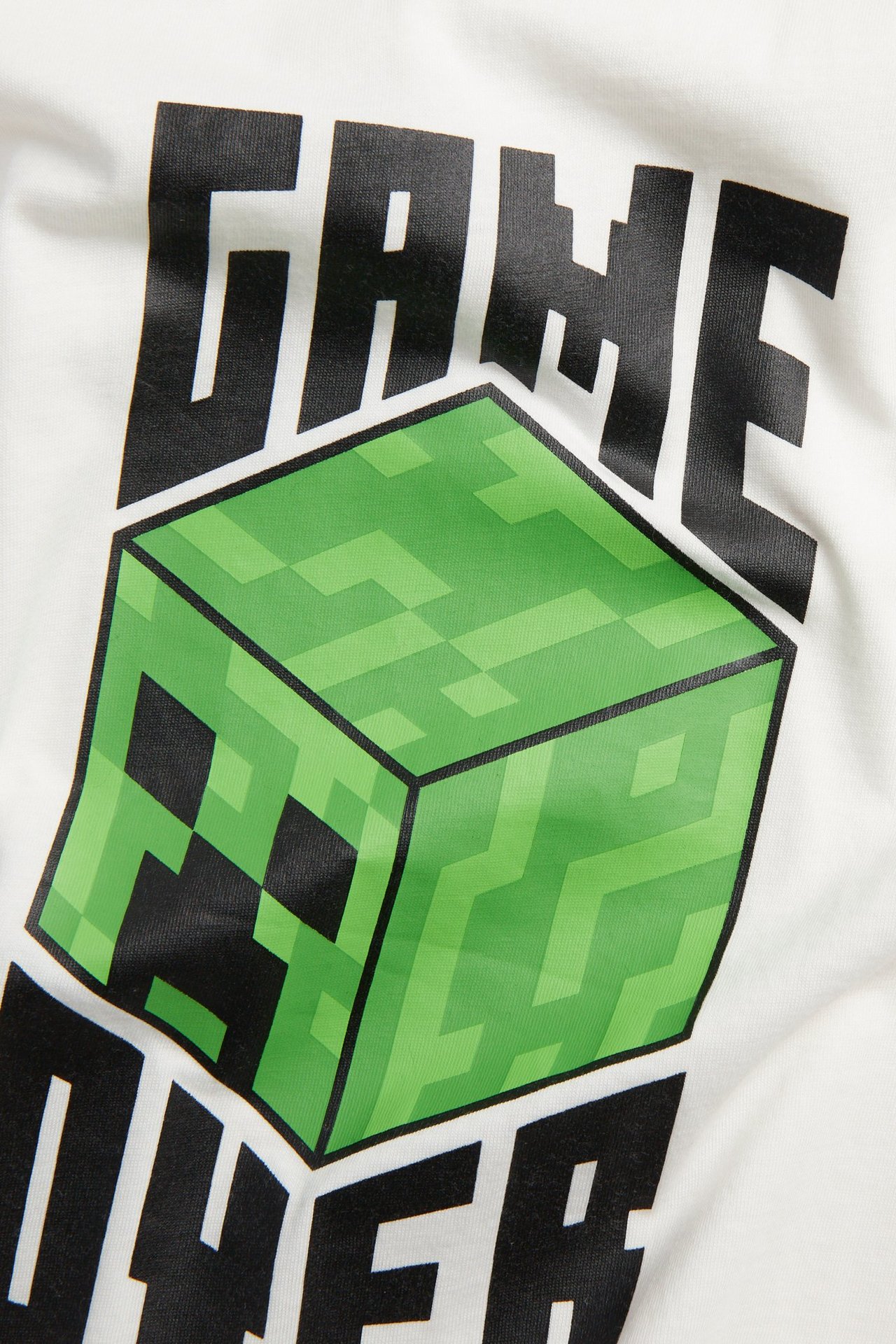 Z bliska: bawełniana koszulka dziecięca w kolorze off-white z zieloną kostką Minecraft i napisem "GAME OVER".