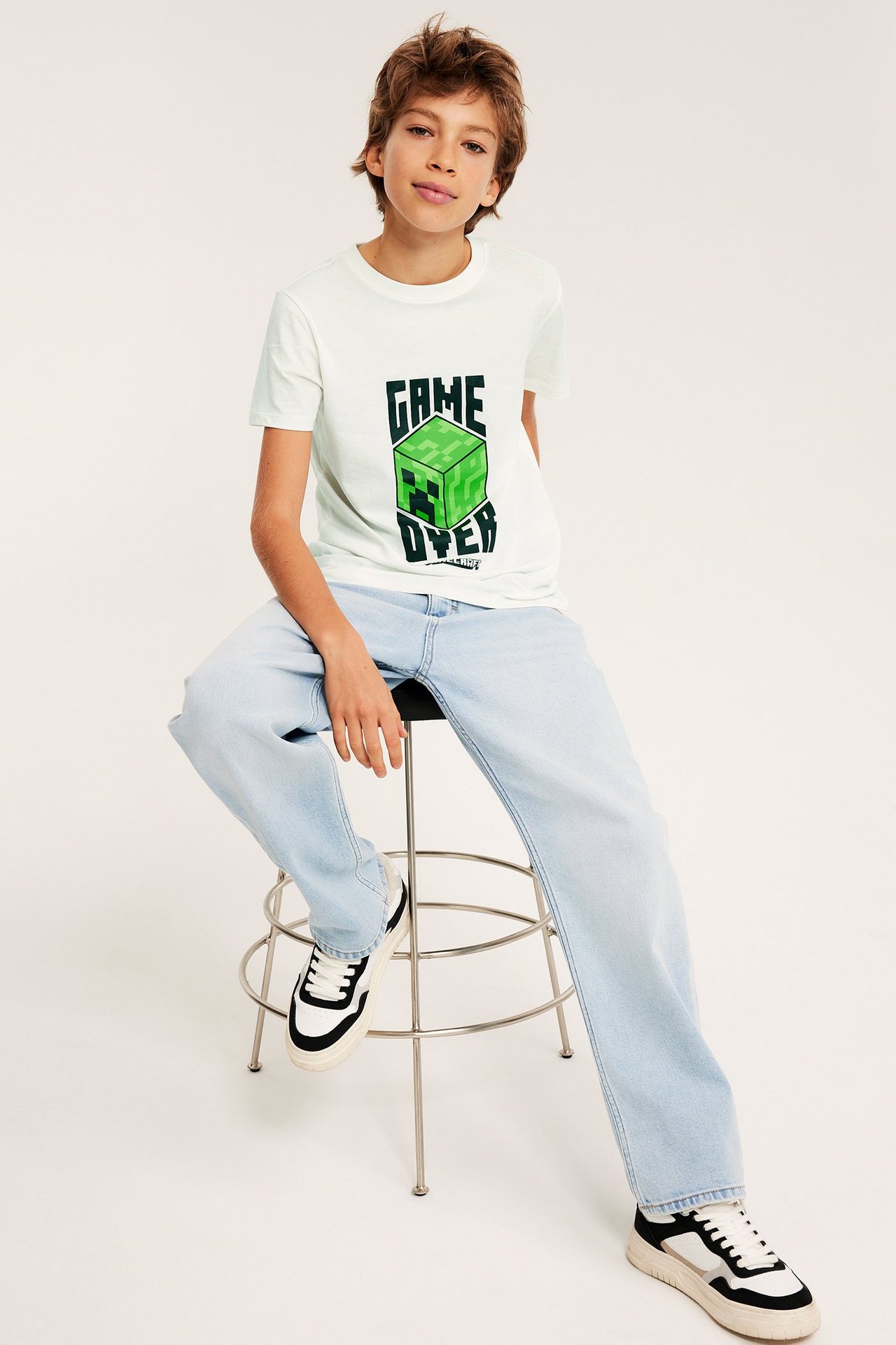 Z przodu: dziecko w białym bawełnianym t-shircie Minecraft, jasnoniebieskich jeansach, czarno-białe sneakersy.