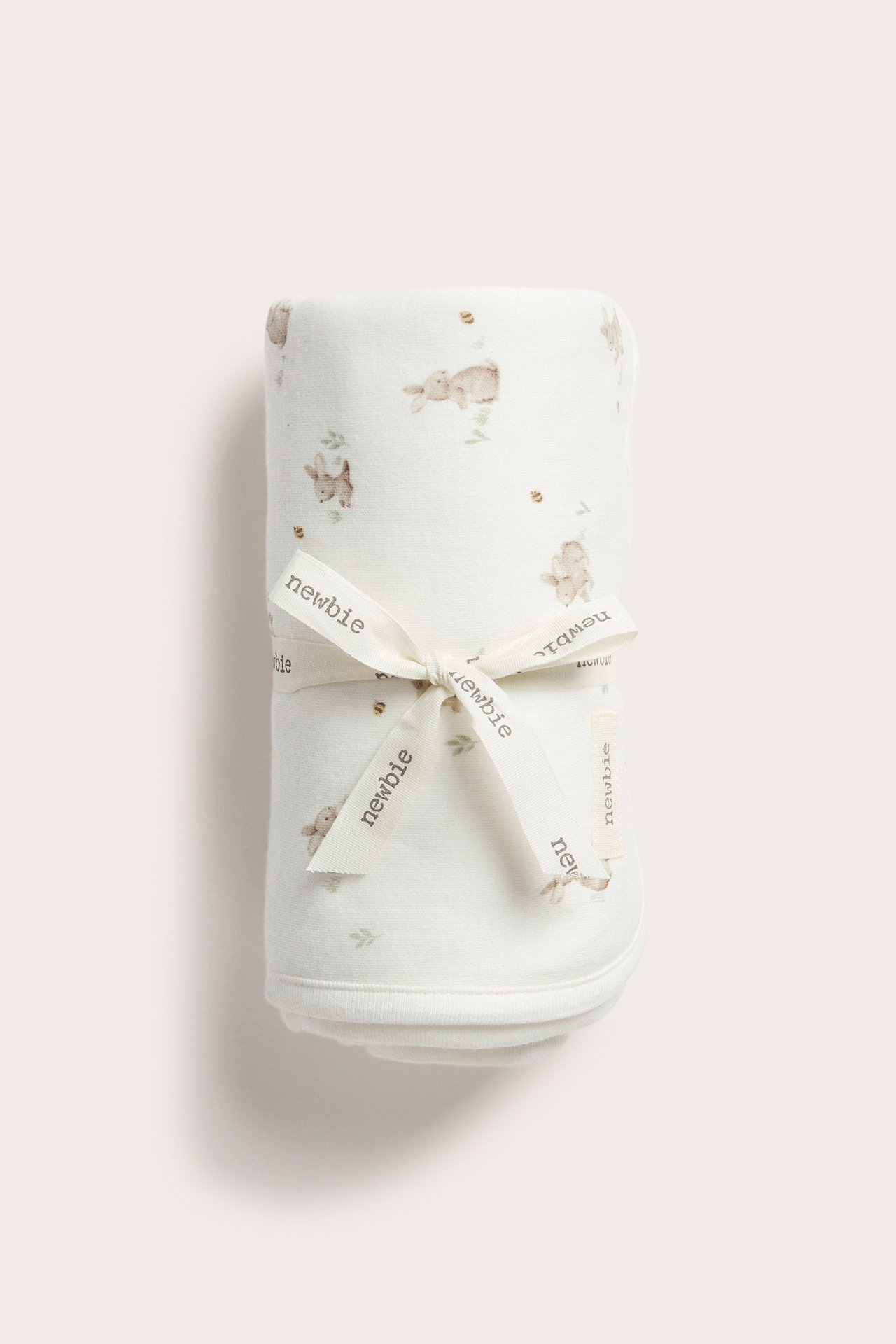 Frontvisning av et offwhite bomulls babyteppe med kaninmønster, rullet og bundet med et Newbie-bånd.