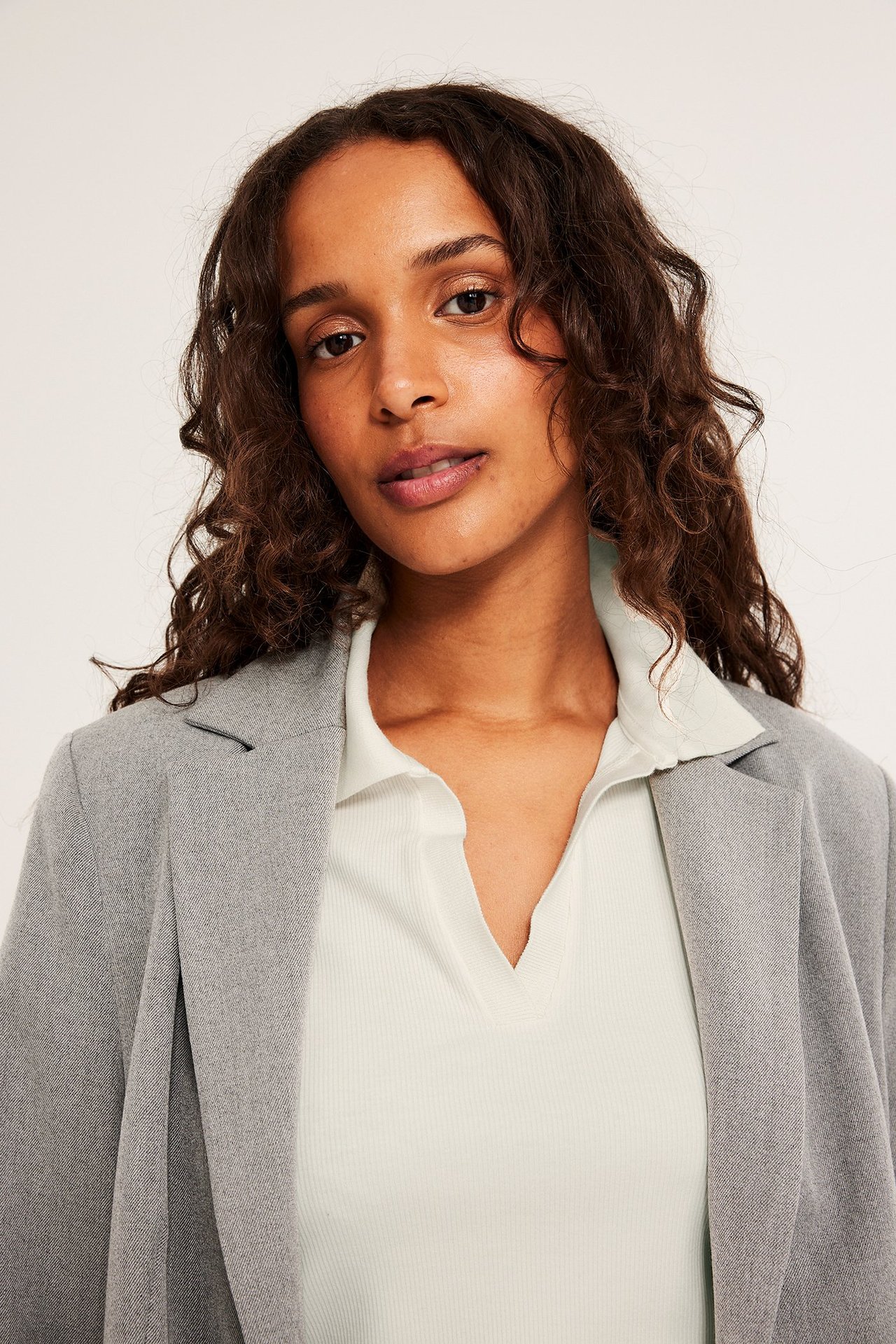 Lähikuva: nainen pitää luonnonvalkoista ribbineulottua lyhythihaista poolopuseroa harmaan bleiserin alla.