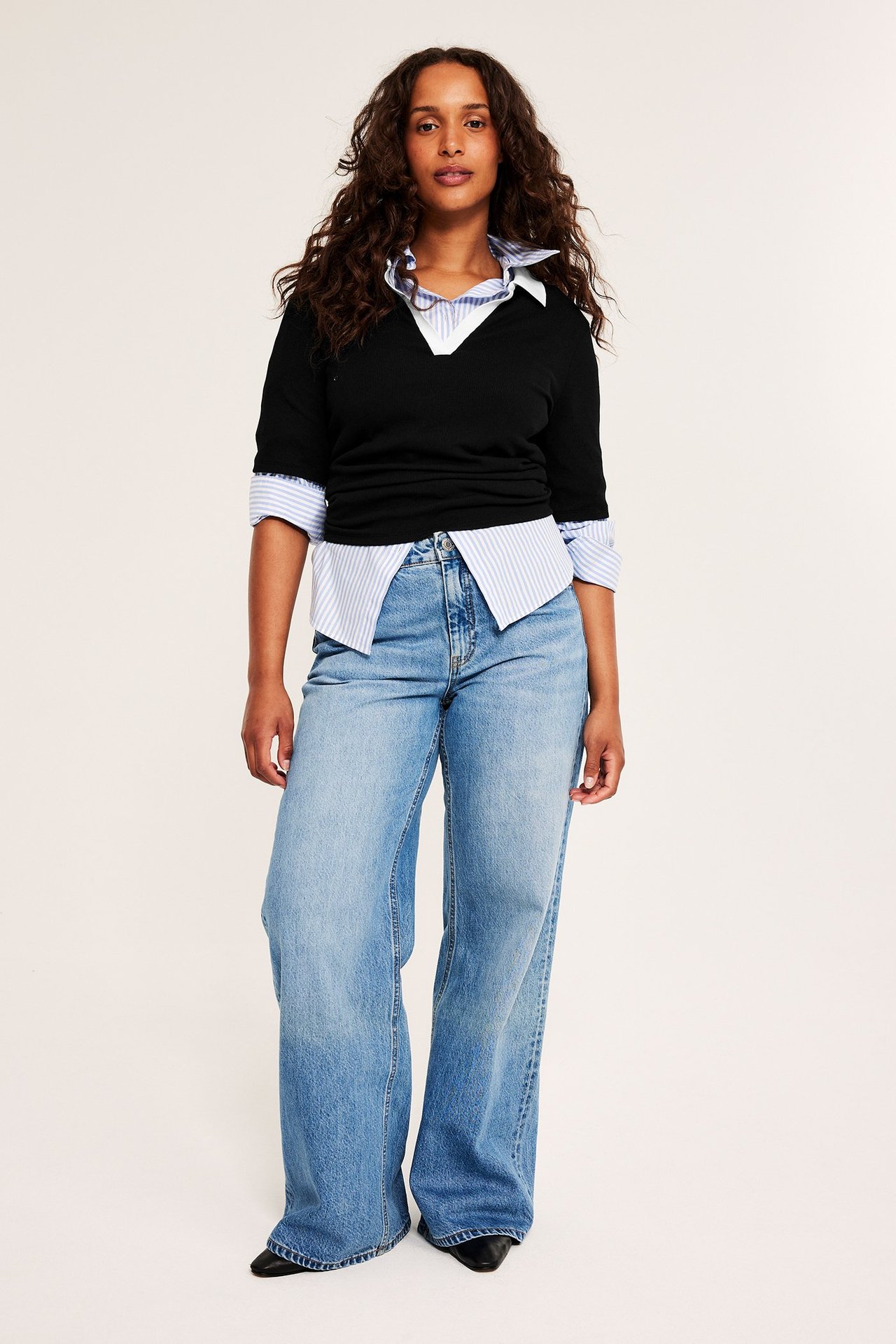 Nainen mustassa V-kaula-aukkoisessa lyhythihaisessa topissa, raidallisessa paidassa, vaaleansinisissä leveissä farkuissa ja mustissa kengissä.