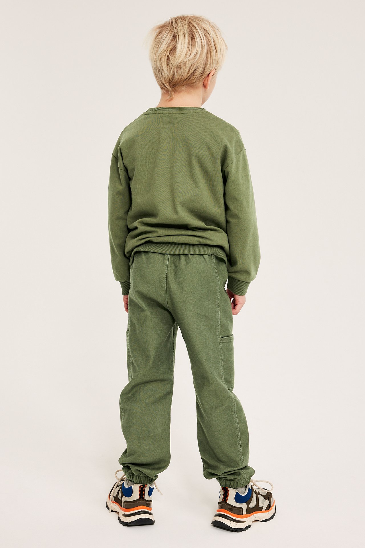 Ryggvisning av et barn i mørkegrønne pull-on bukser med lommer, en matchende genser og joggesko.