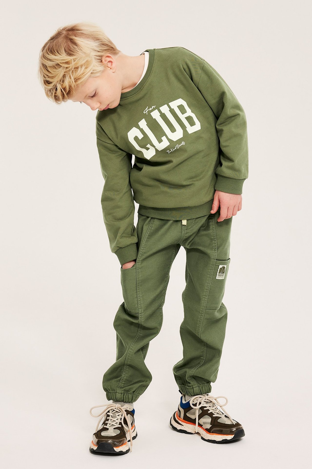 Framifrån, ett barn i mörkgröna pull-on cargobyxor med sidfickor, en matchande 'CLUB' tröja och sneakers.