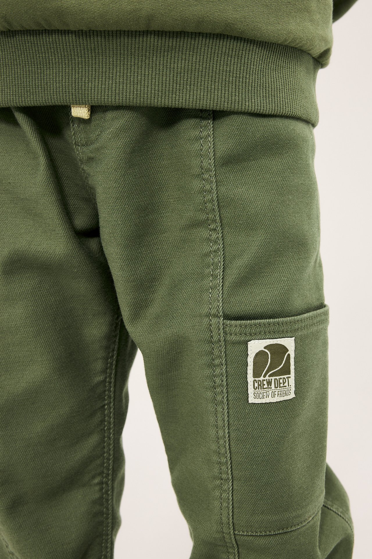 Nærbilde av mørkegrønn bukse til barn med elastisk midje, snøring og sidelommeetikett.