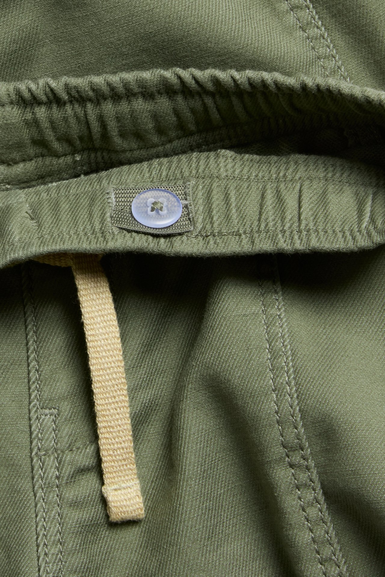 Nærbilde av mørkegrønne pull-on bukser med elastisk linning, lyseblå knapp og beige snøring.