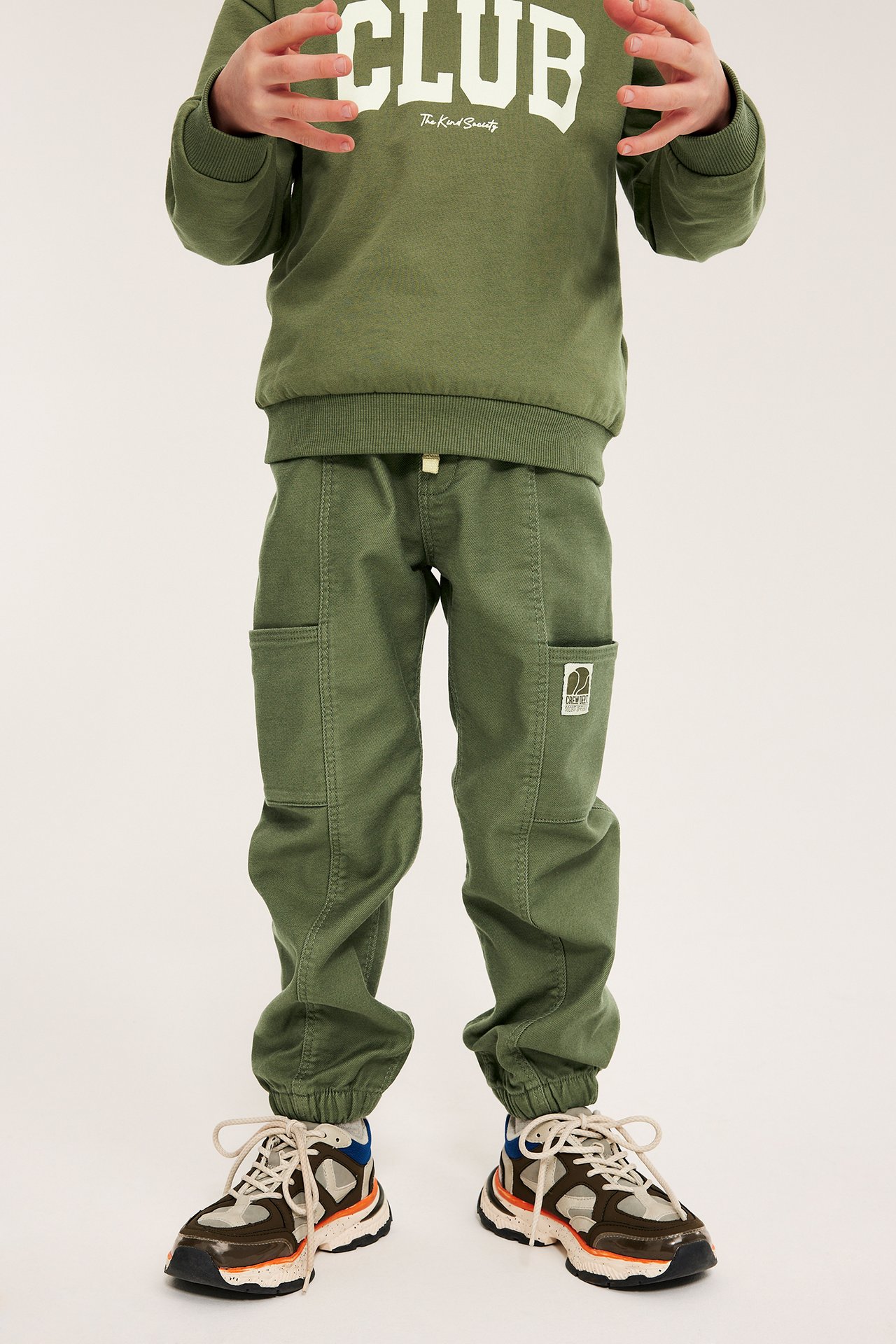 Frontvy av ett barn i mörkgröna cargobyxor med resår, en olivgrön tröja med tryck och sneakers.