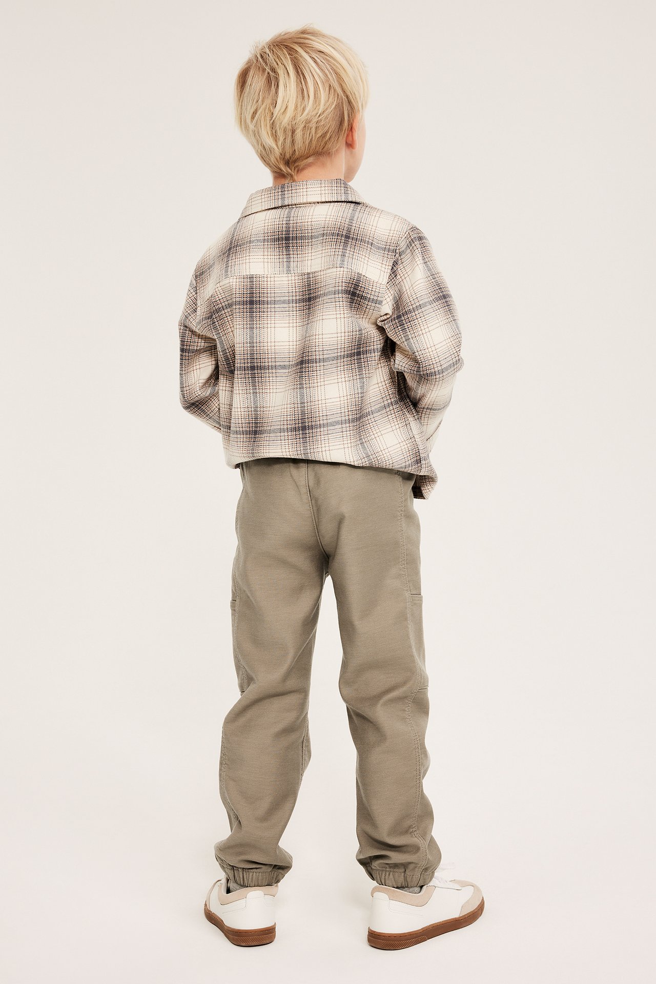 Bakfra sett, et barn i beige bukser med mansjetter, rutete skjorte og hvite joggesko.