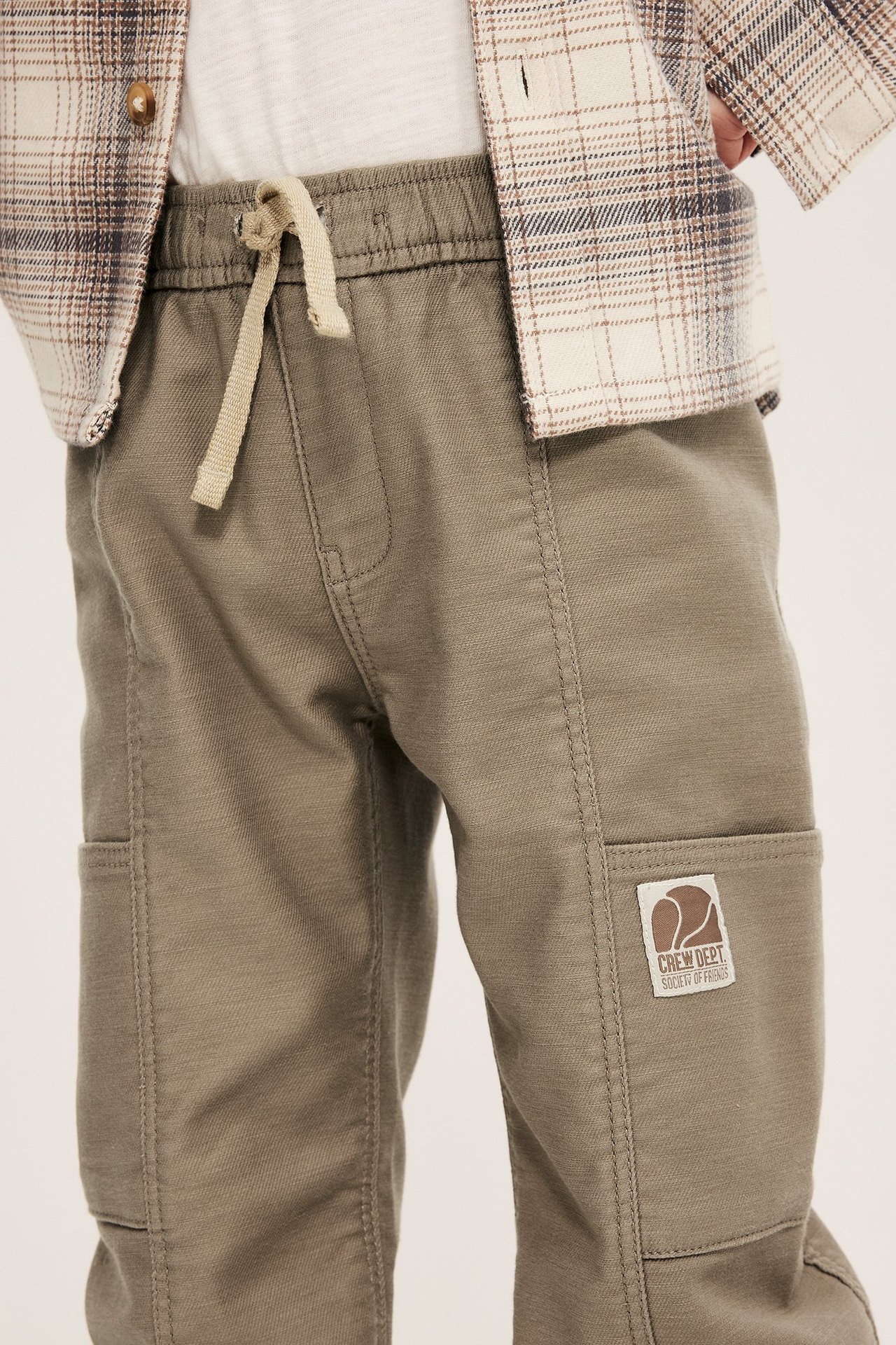 Närbild på ett barn i beiga pull-on byxor med dragsko, vit t-shirt och öppen rutig skjorta.