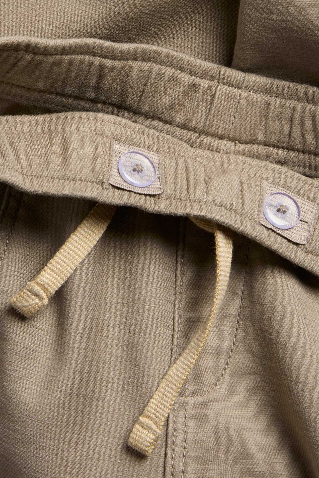 Zbliżenie na beżowe spodnie typu pull-on z elastycznym pasem, beżowym sznurkiem i guzikami do regulacji.