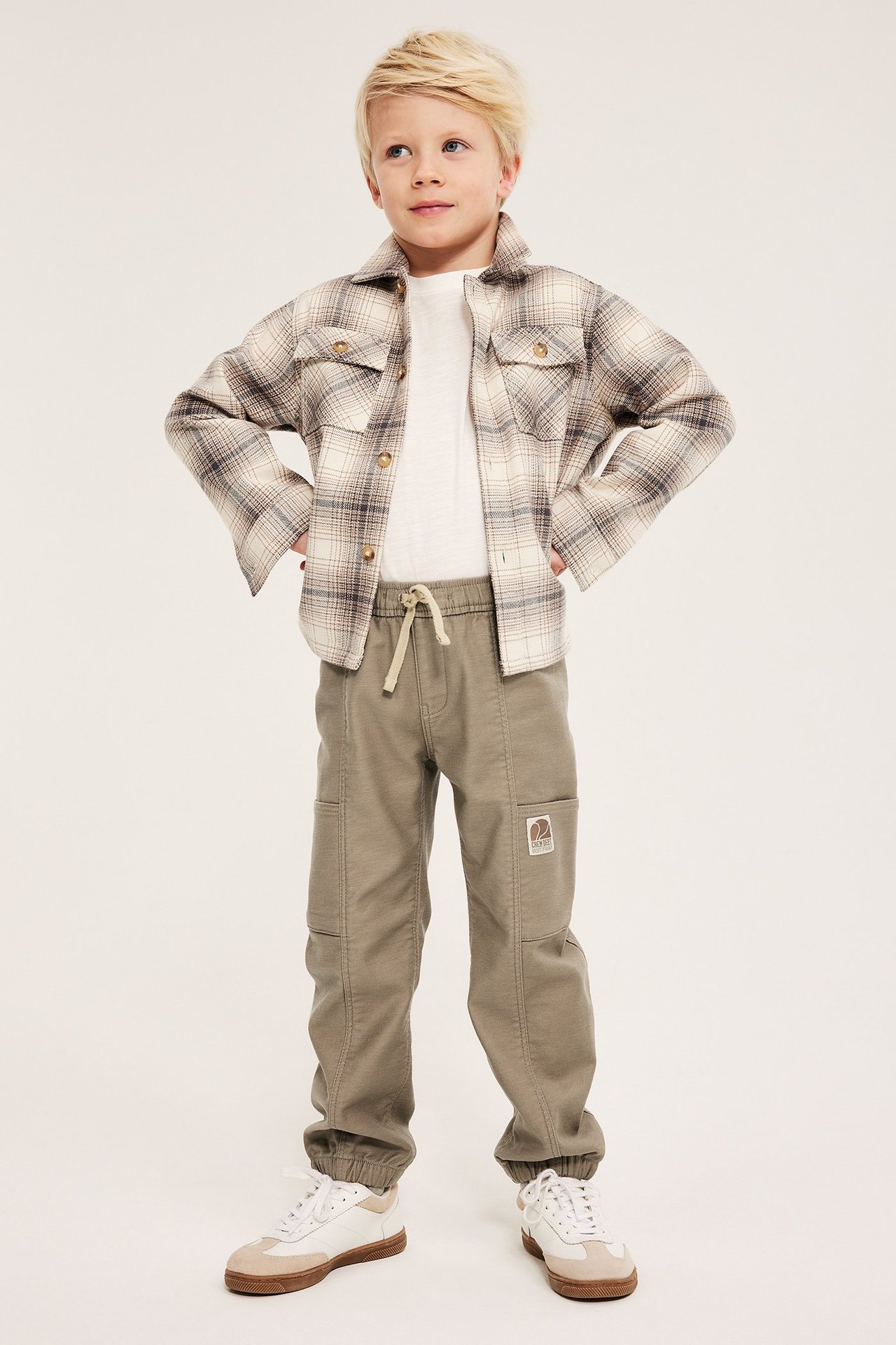Frontvy av ett barn i beiga pull-on cargobyxor, vit t-shirt, rutig shacket och sneakers.