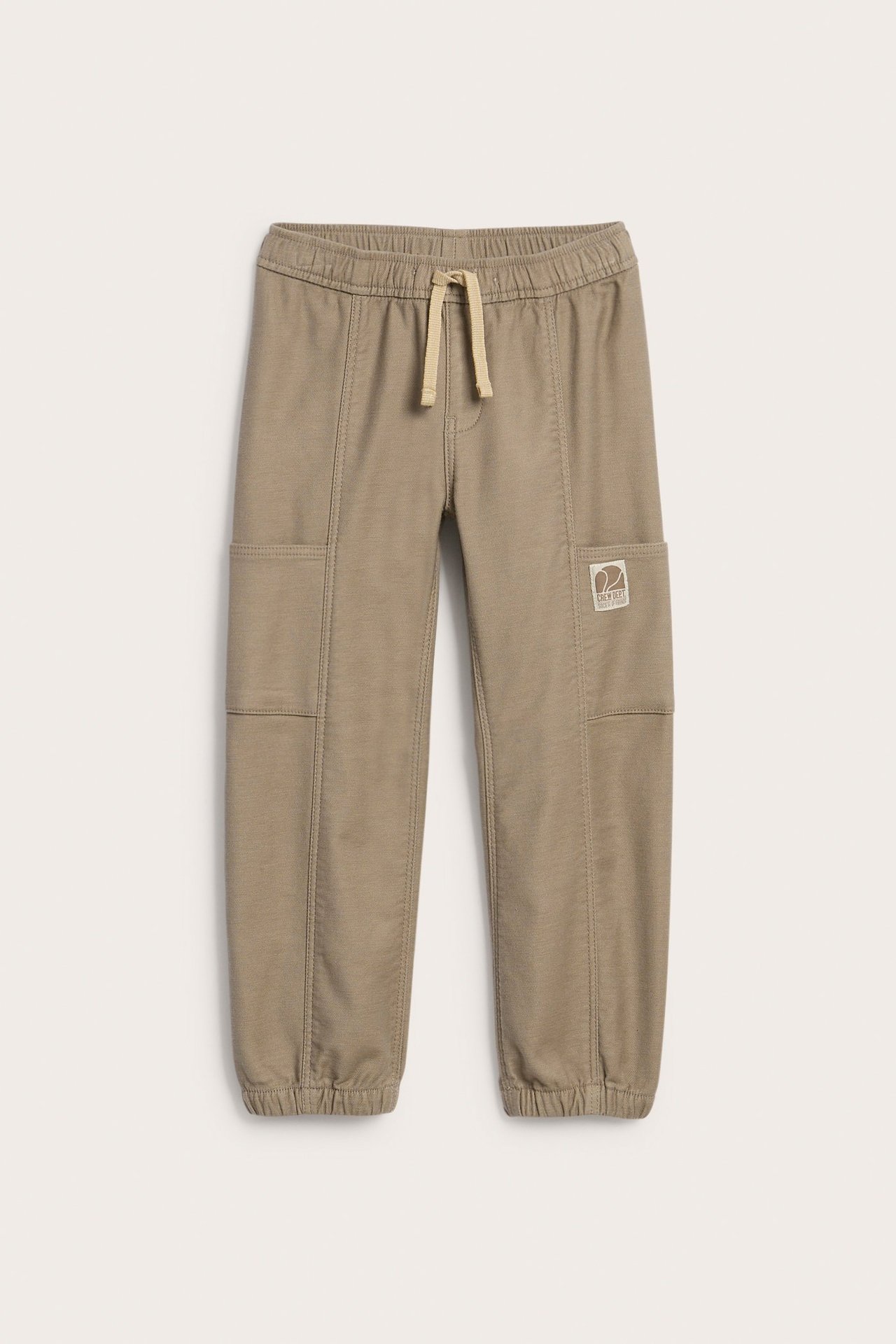 Framifrån, beiga barnbyxor i bomullsblandning med resårmidja, dragsko, cargofickor och ribbade muddar.