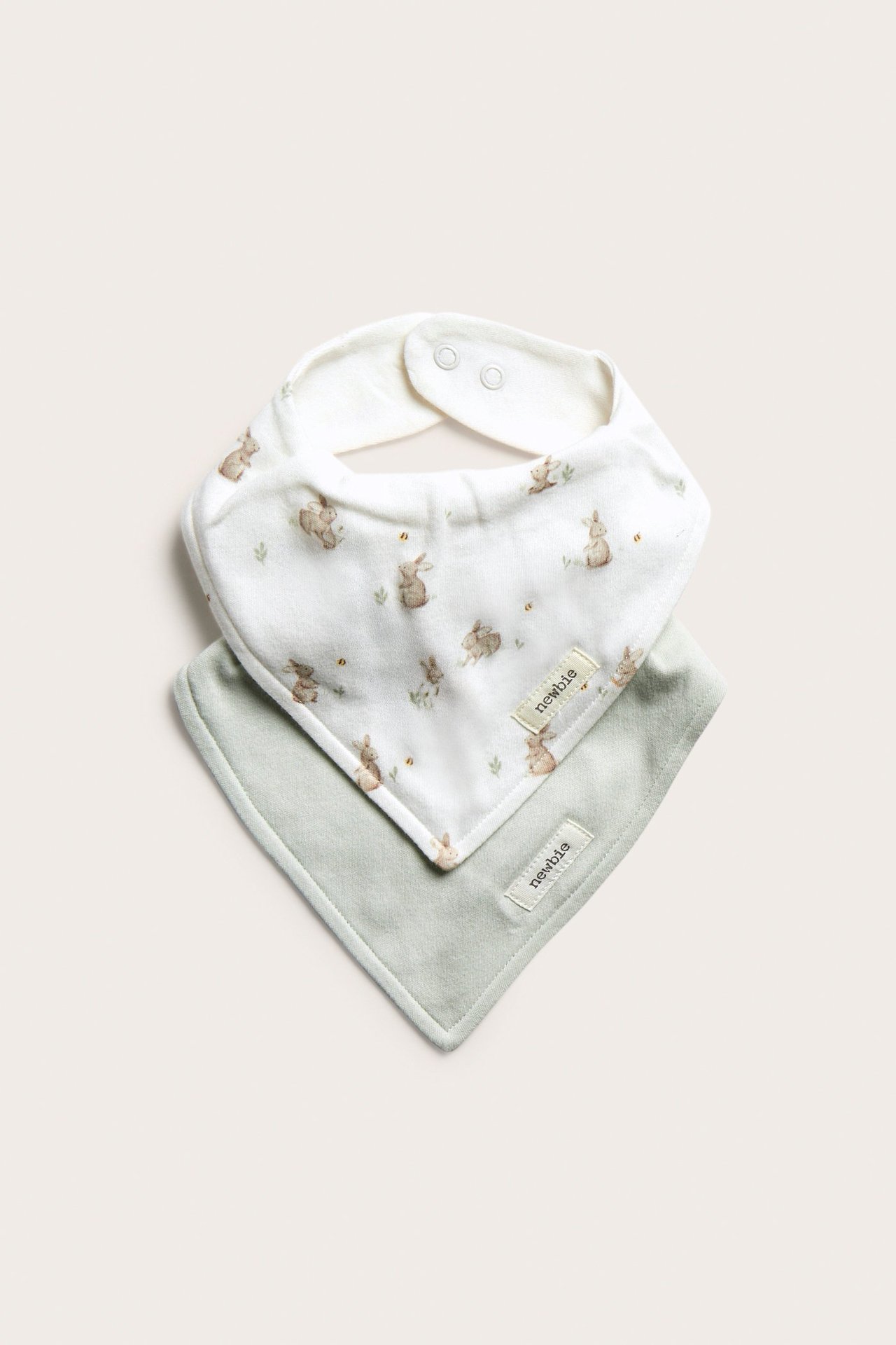 Widok z przodu: Dwa bawełniane śliniaki: jeden ecru z nadrukiem królików, jeden jasnozielony, oba z zatrzaskami.