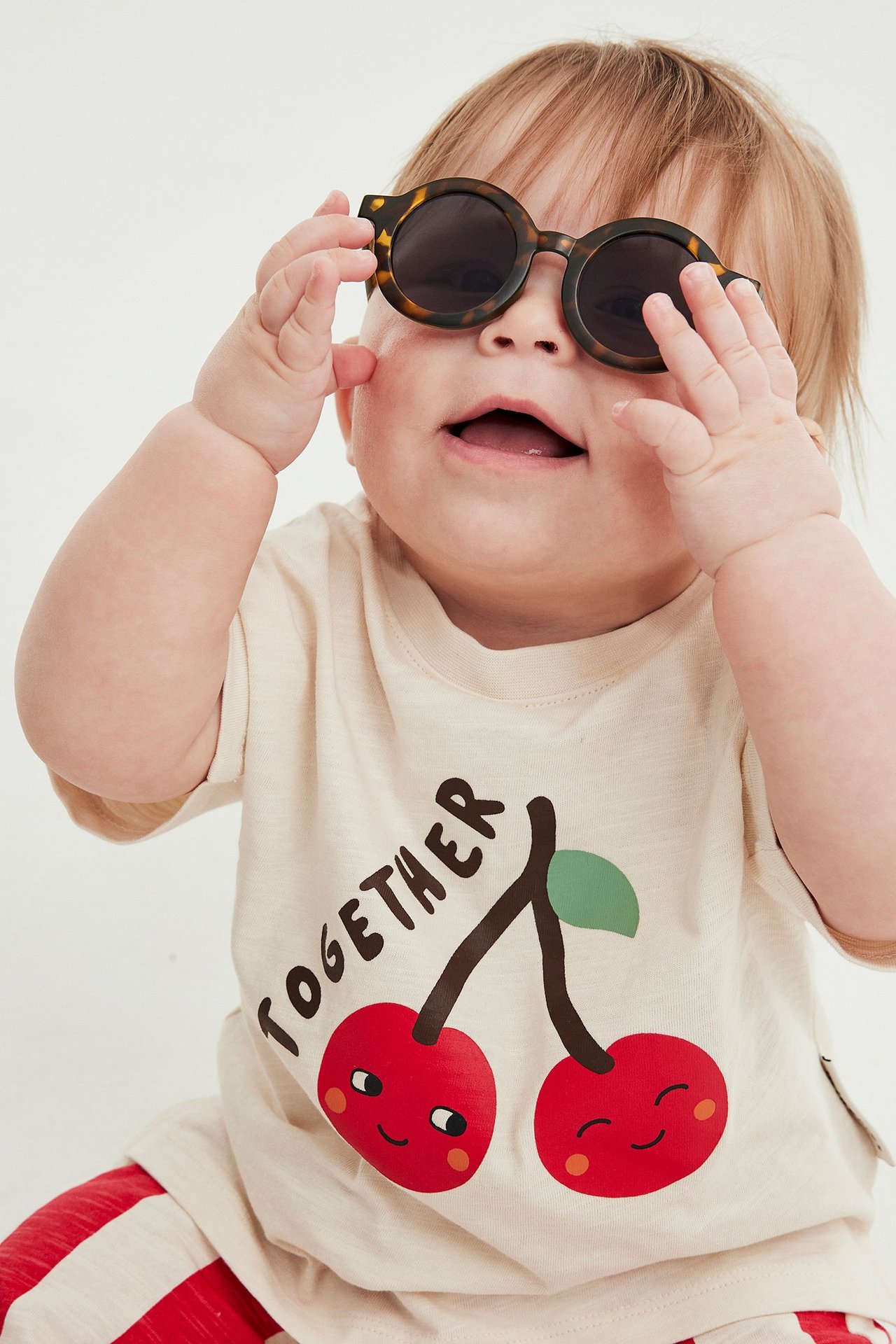 Niemowlę ma na sobie okrągłe wzorzyste brązowe okulary przeciwsłoneczne i jasnobeżowy t-shirt z nadrukiem wiśni.