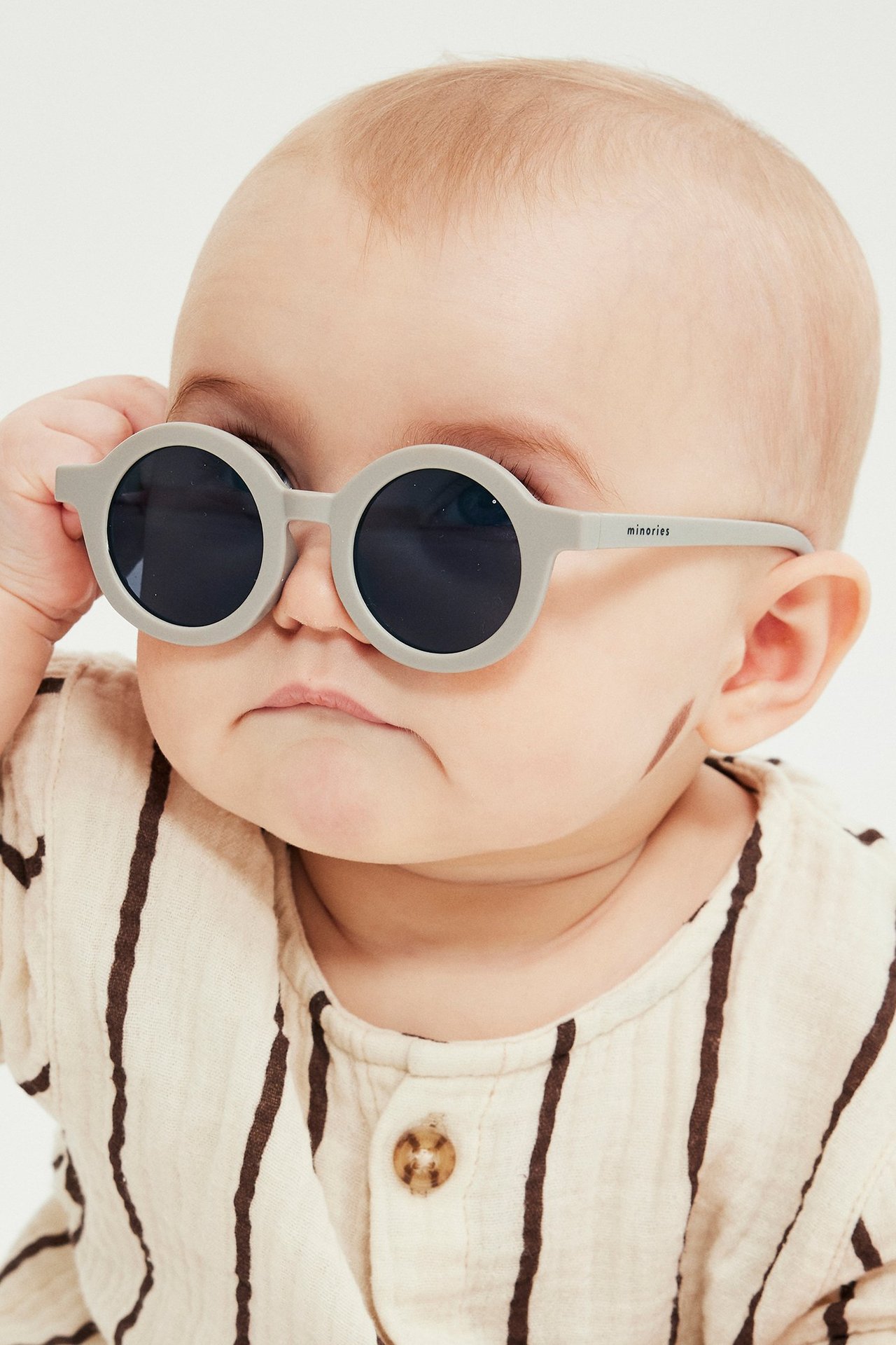 Zbliżenie, widok z przodu, dziecka poprawiającego okrągłe beżowe okulary przeciwsłoneczne. Dziecko ma jasny pasiasty top.