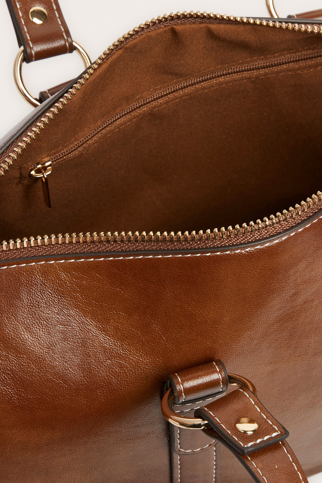Nærbilde av en åpen brun håndveske med gyllen glidelås, brunt fôr og hvite kontrastsømmer.