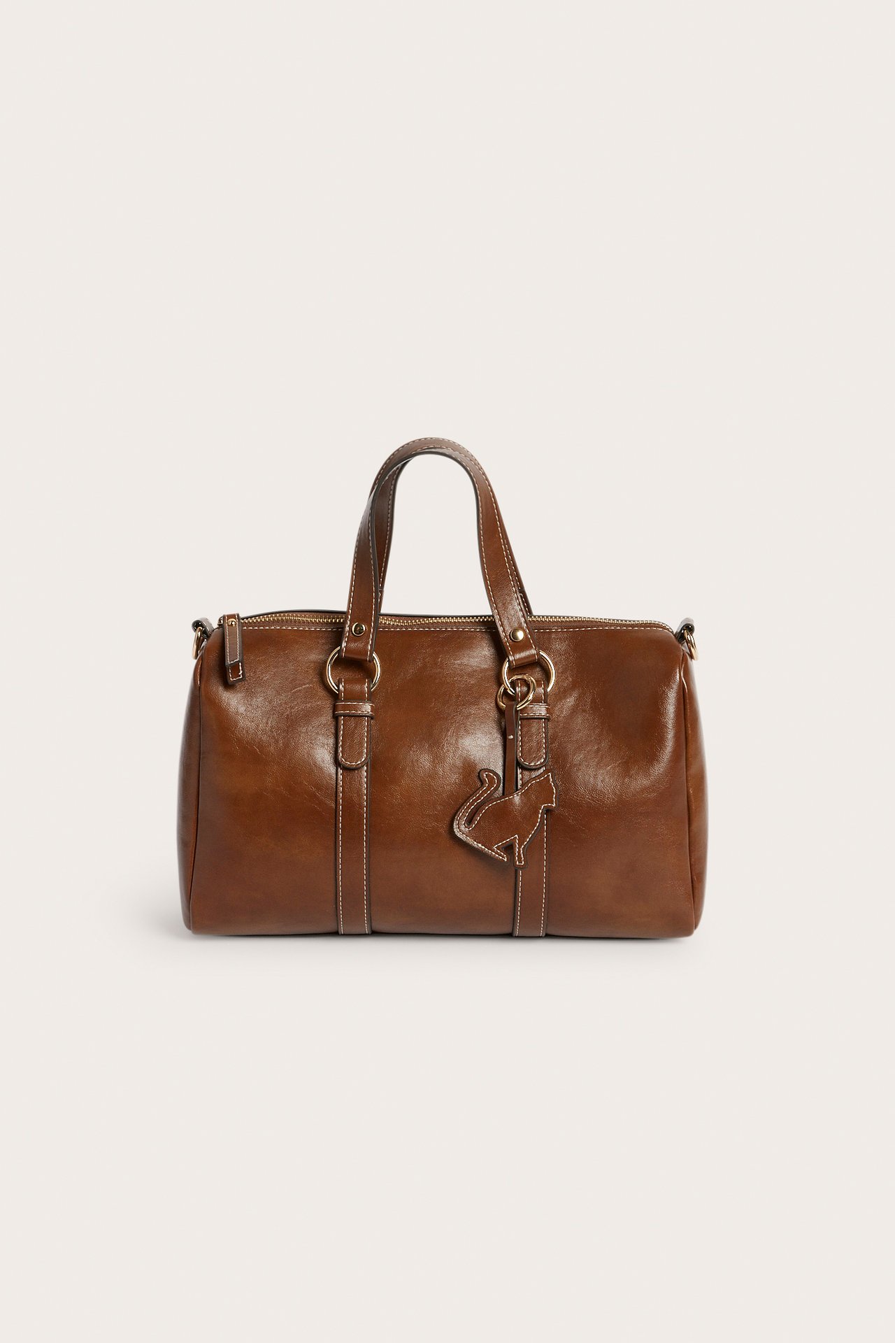 Frontvisning av en brun håndveske med håndtak, glidelås og et dekorativt katteanheng.