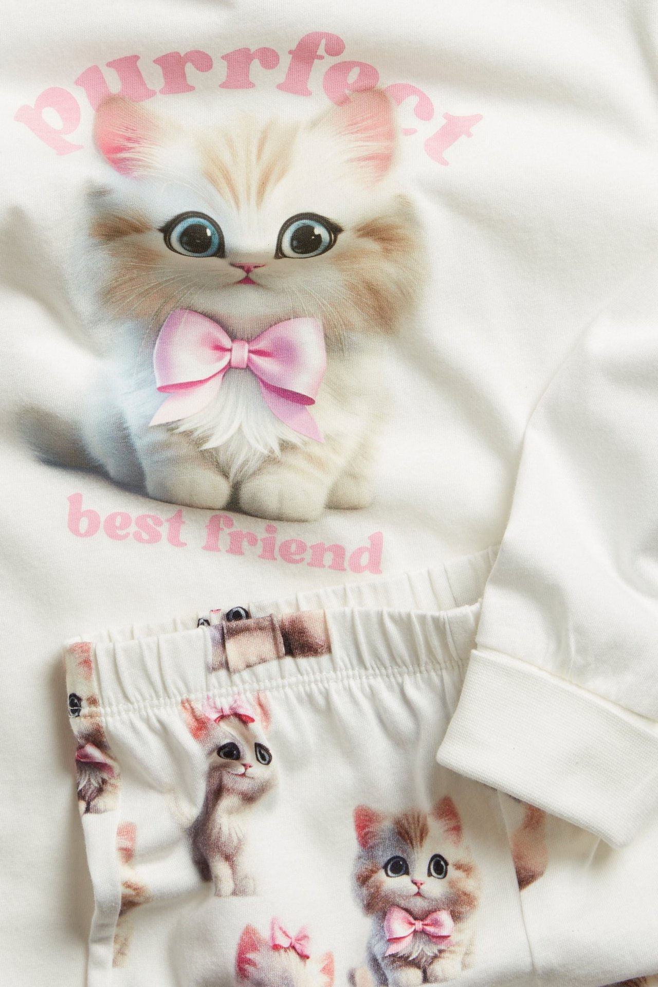 Luonnonvalkoiset puuvillapyjamat. Yläosassa suuri kissaprintti, housuissa kissakuvio.
