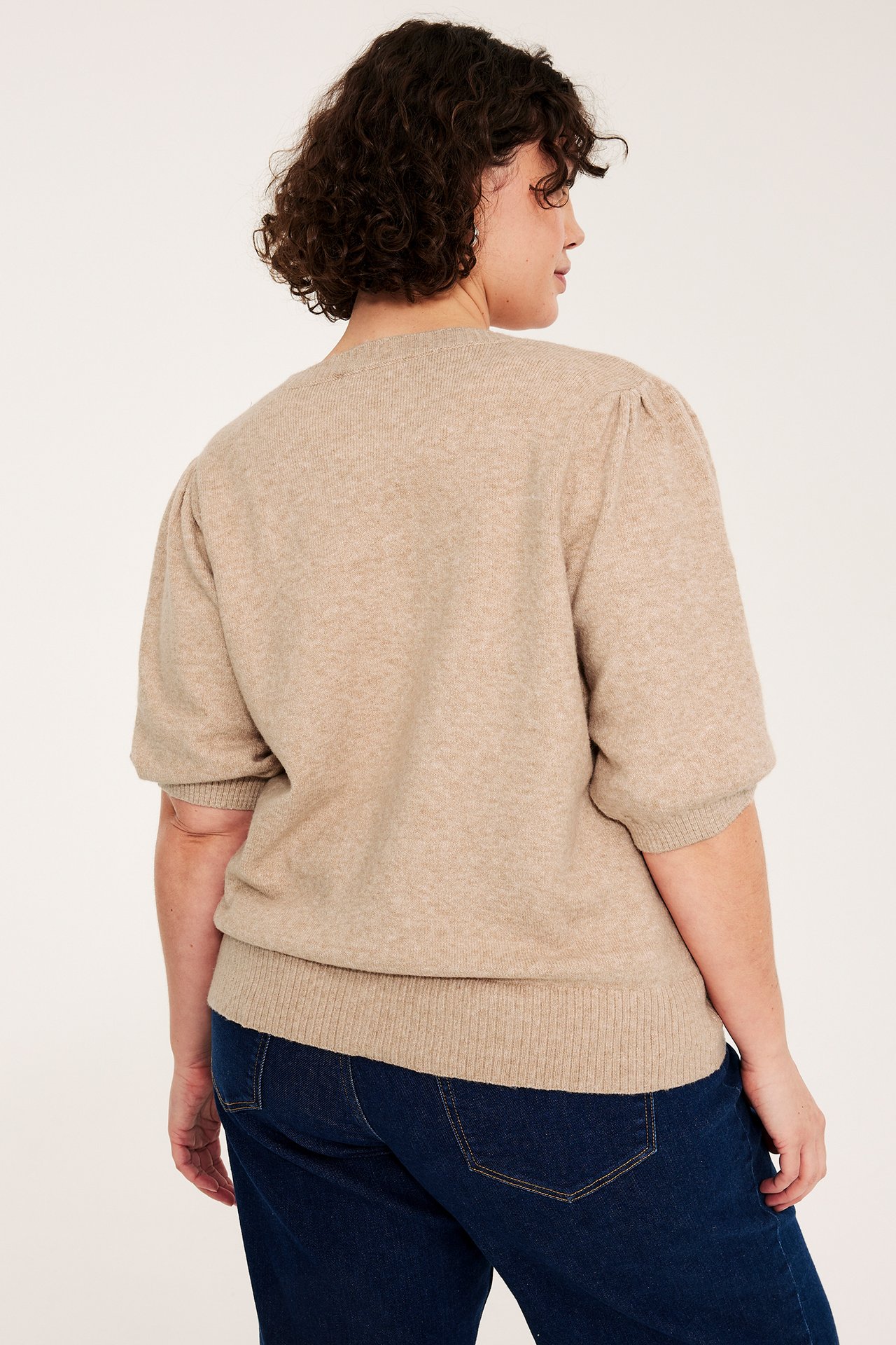 Nainen takaa: yllään beige melange lyhythihainen neuletakki pussihihoilla ja tummansiniset farkut.