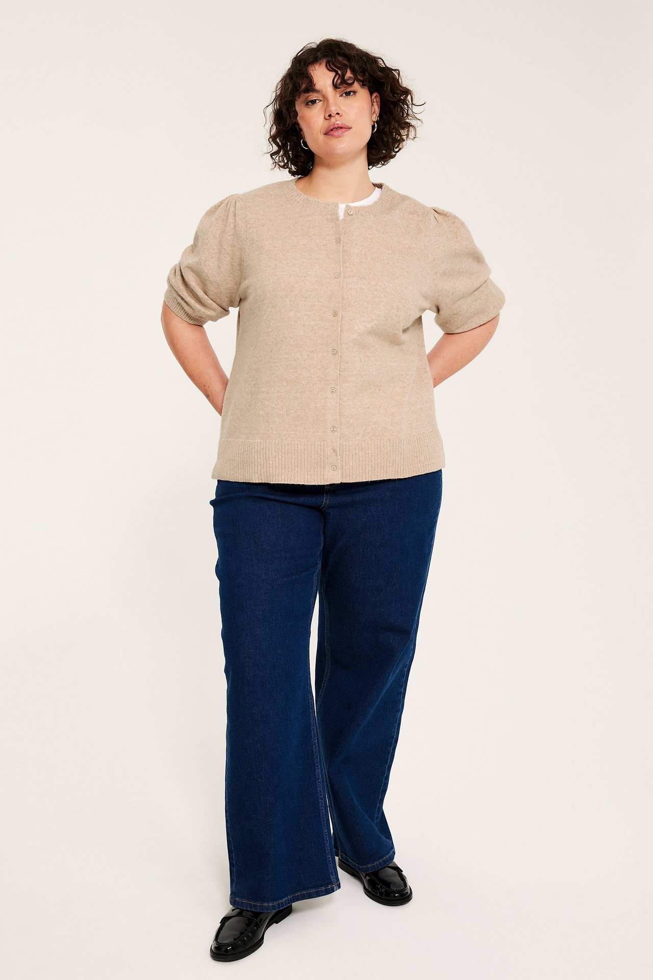 Etunäkymä: Nainen meleratussa beigessä puffihihaisessa neuletakissa, valkoisessa t-paidassa, tummansinisissä farkuissa ja mustissa loafer-kengissä.
