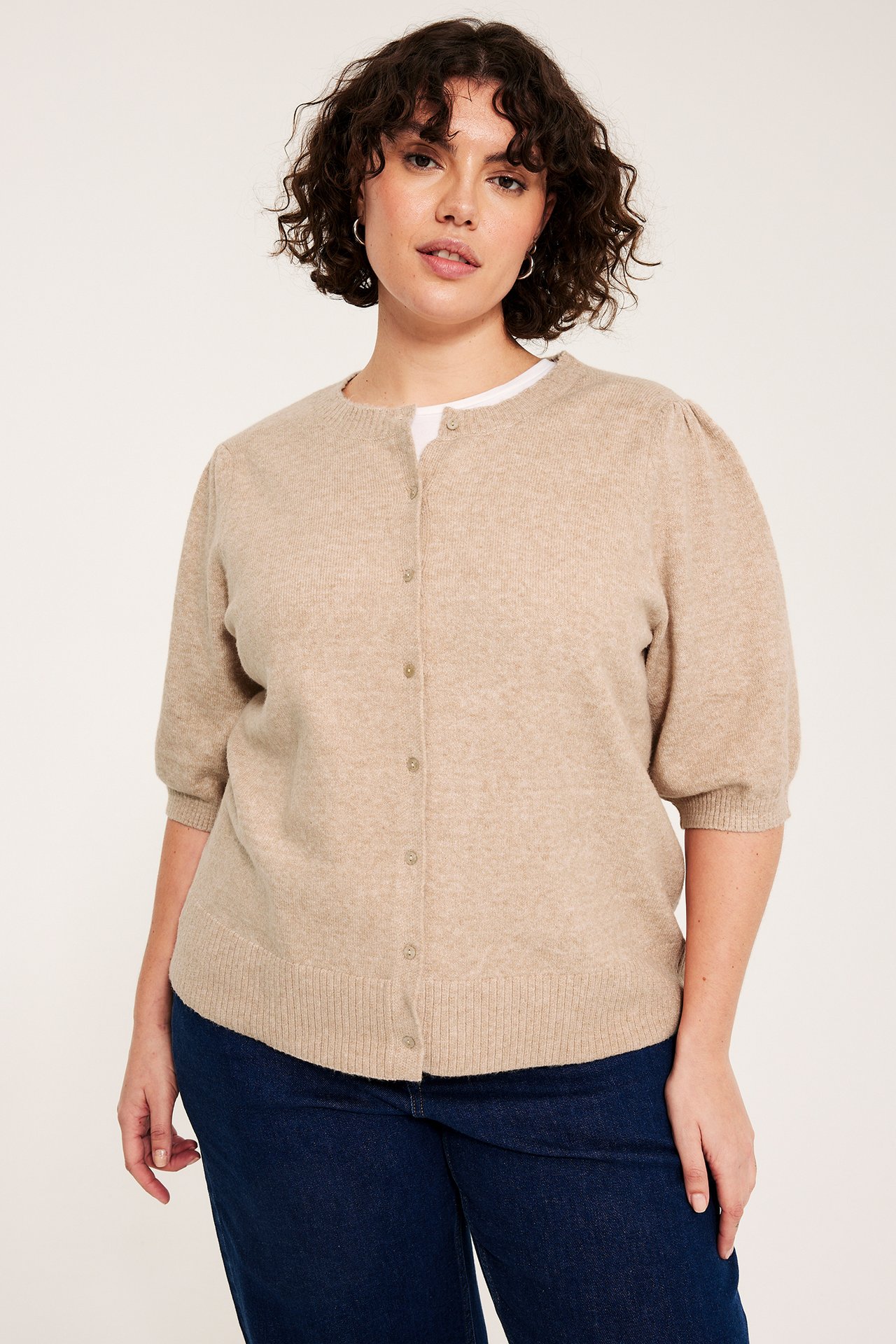 Edestä: Nainen beigessä meleeratussa lyhythihaisessa pussihihaisessa neuletakissa, valkoinen toppi, tummansiniset farkut.