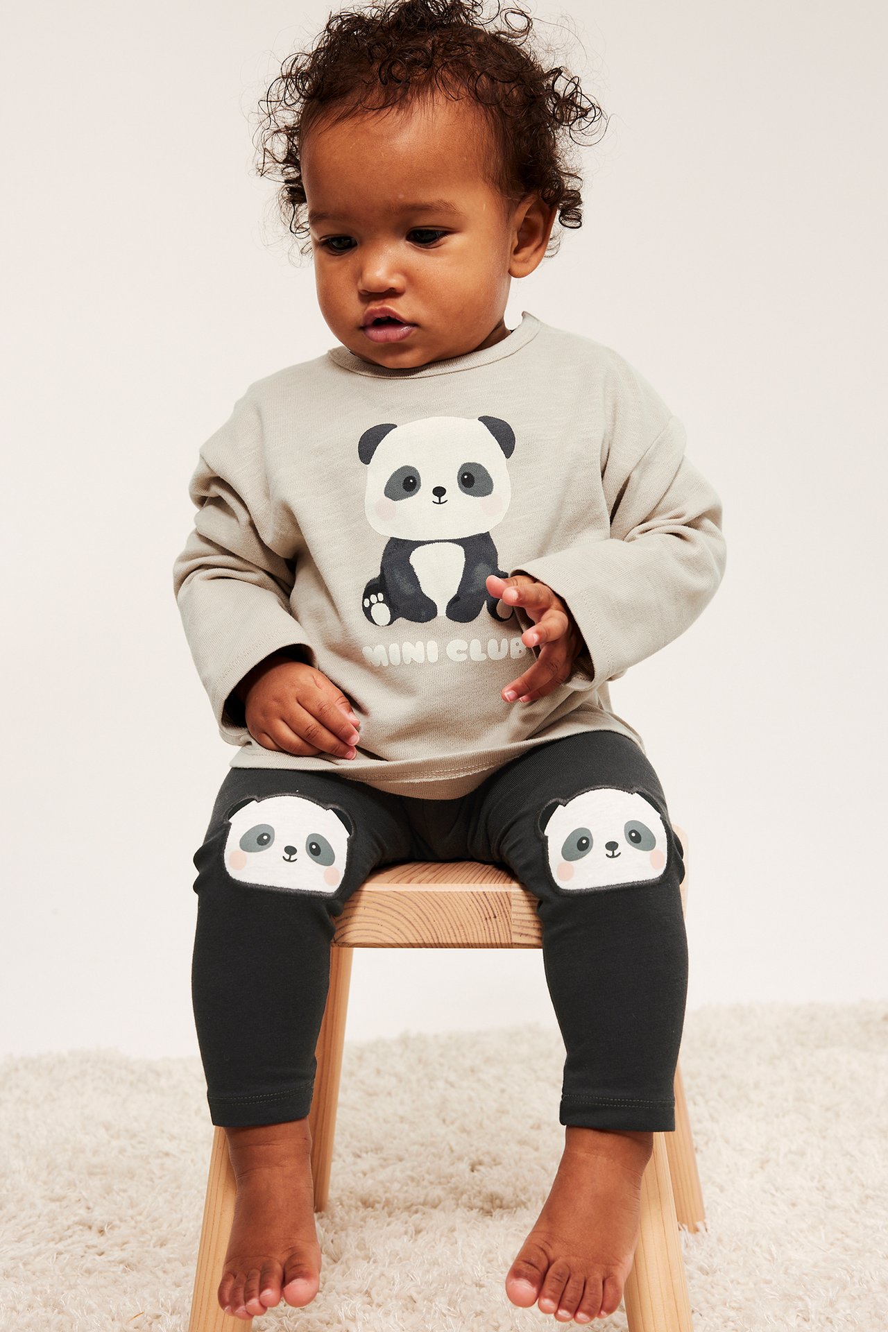 Widok z przodu: niemowlę w czarnych legginsach z aplikacjami pandy na kolanach i jasnoszarej bluzce z pandą.