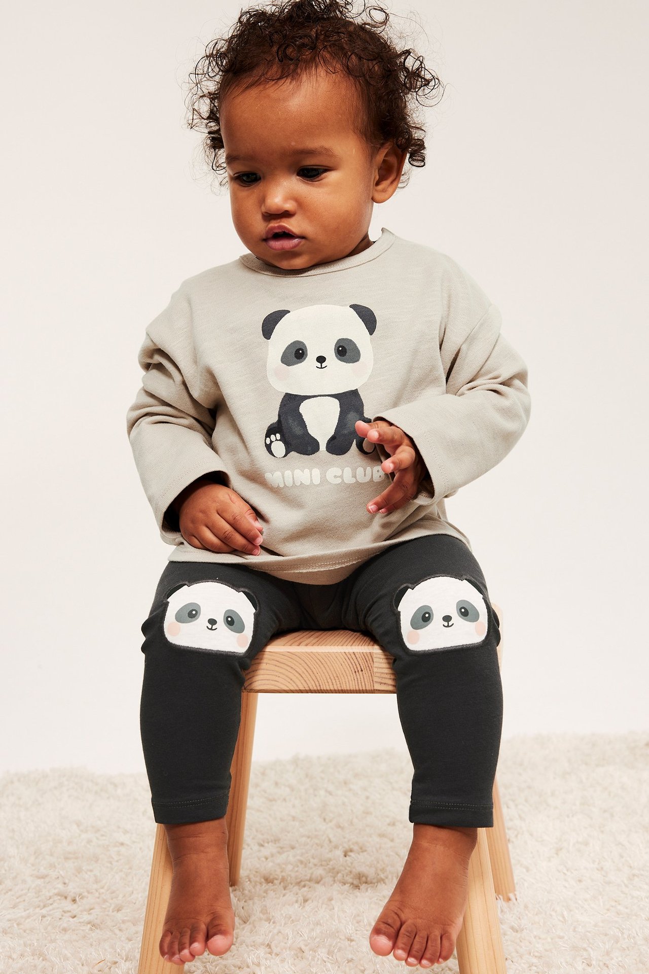 Widok z przodu: niemowlę w czarnych legginsach z aplikacjami pandy na kolanach i jasnoszarej bluzce z pandą.
