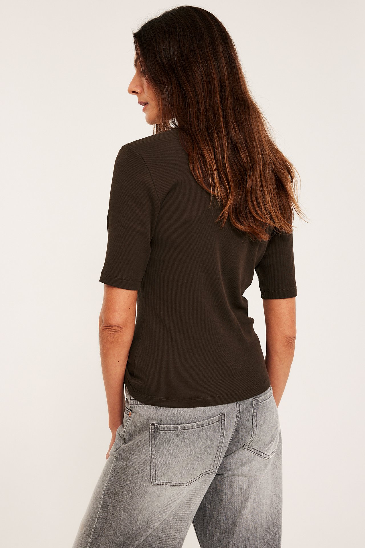 Nainen takaa/sivulta: tummanruskea ribattu lyhythihainen toppi ja vaaleanharmaat farkut.