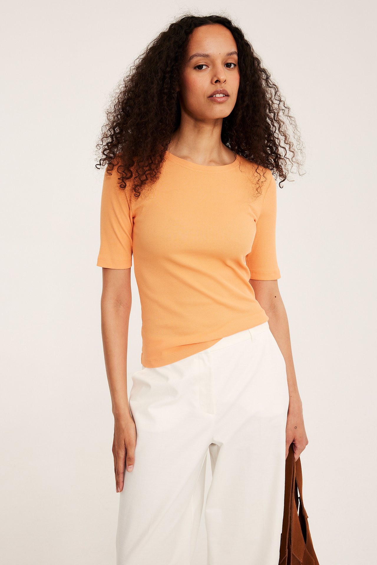 Framifrån: kvinna i orange ribbad kortärmad topp, vita vida byxor och med en brun flätad väska.