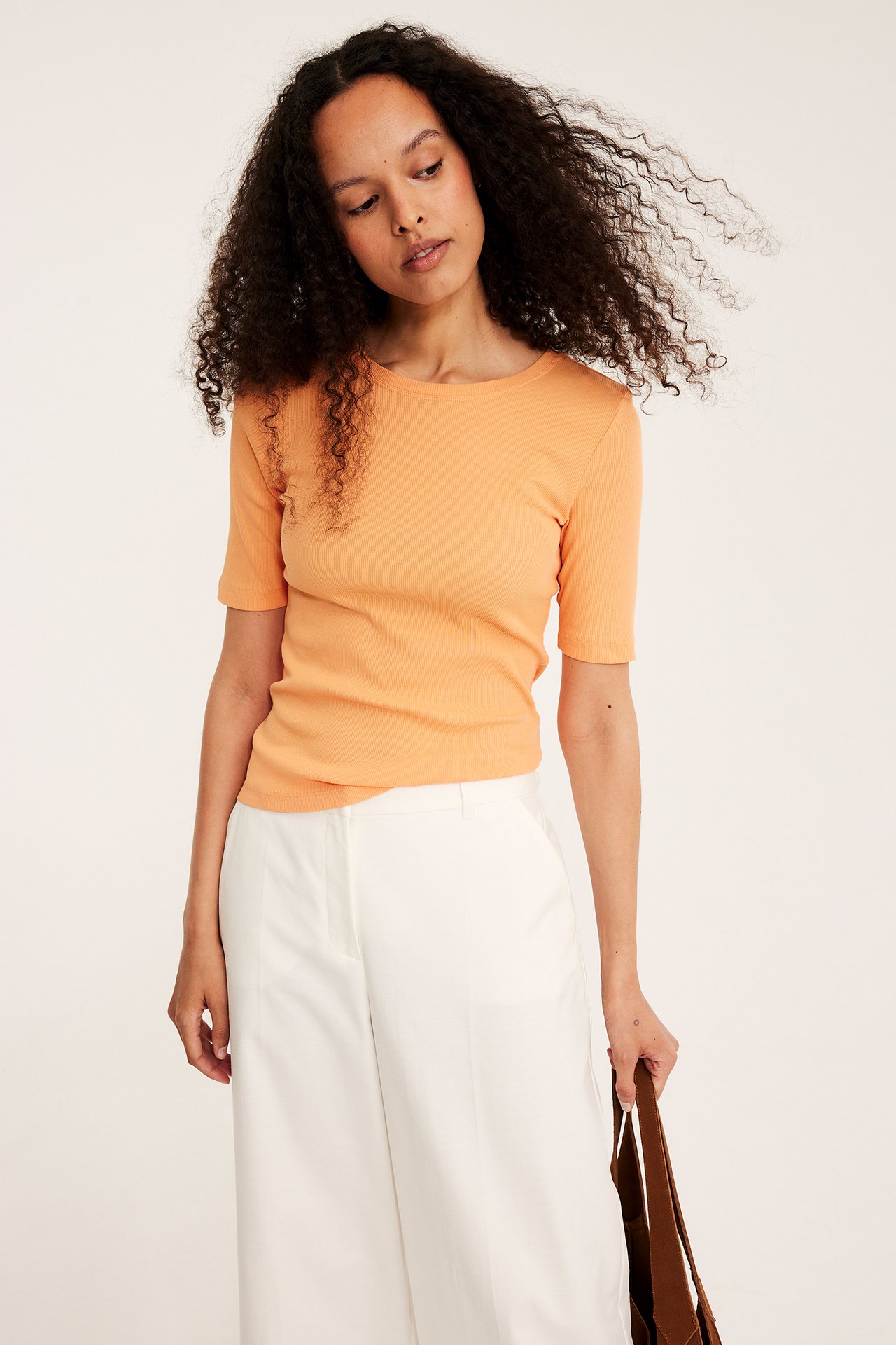 Framifrån: kvinna i orange ribbad kortärmad topp, vita vida byxor, håller en brun väska.