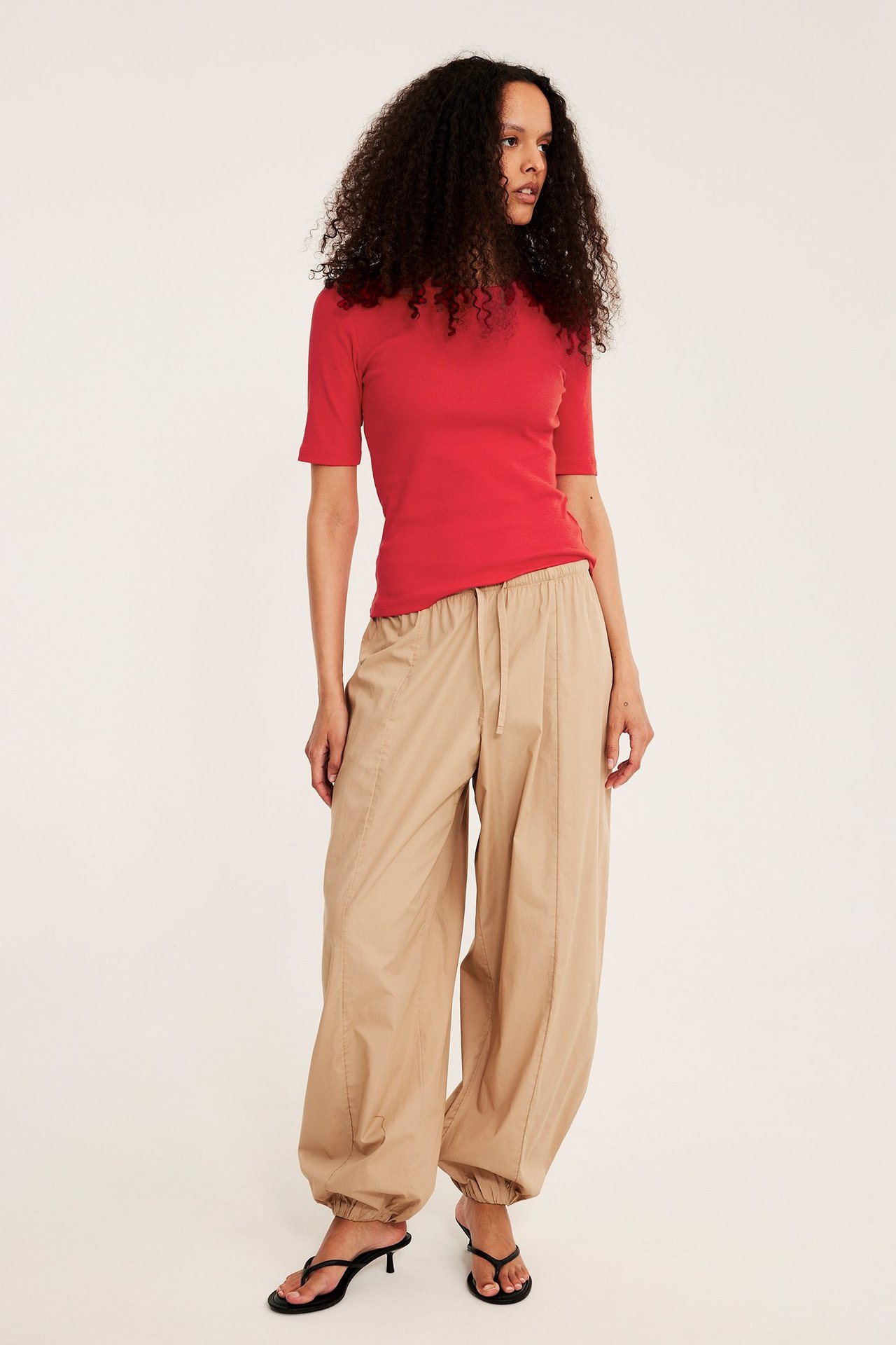Widok z przodu: kobieta w czerwonym prążkowanym topie, beżowych spodniach ze sznurkiem i czarnych sandałach.