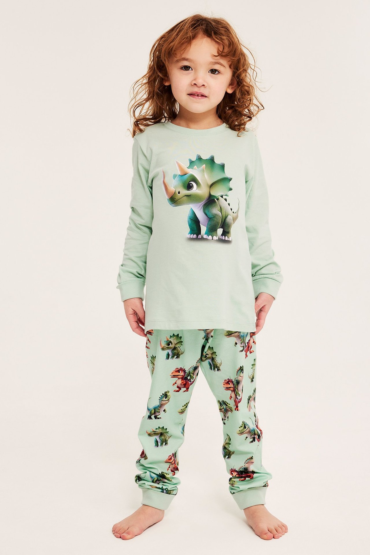 Forfra: Barn iført lysgrønn bomullspyjamas. Topp med stort dinosaurtrykk, bukser med heldekkende dinosauermønster.