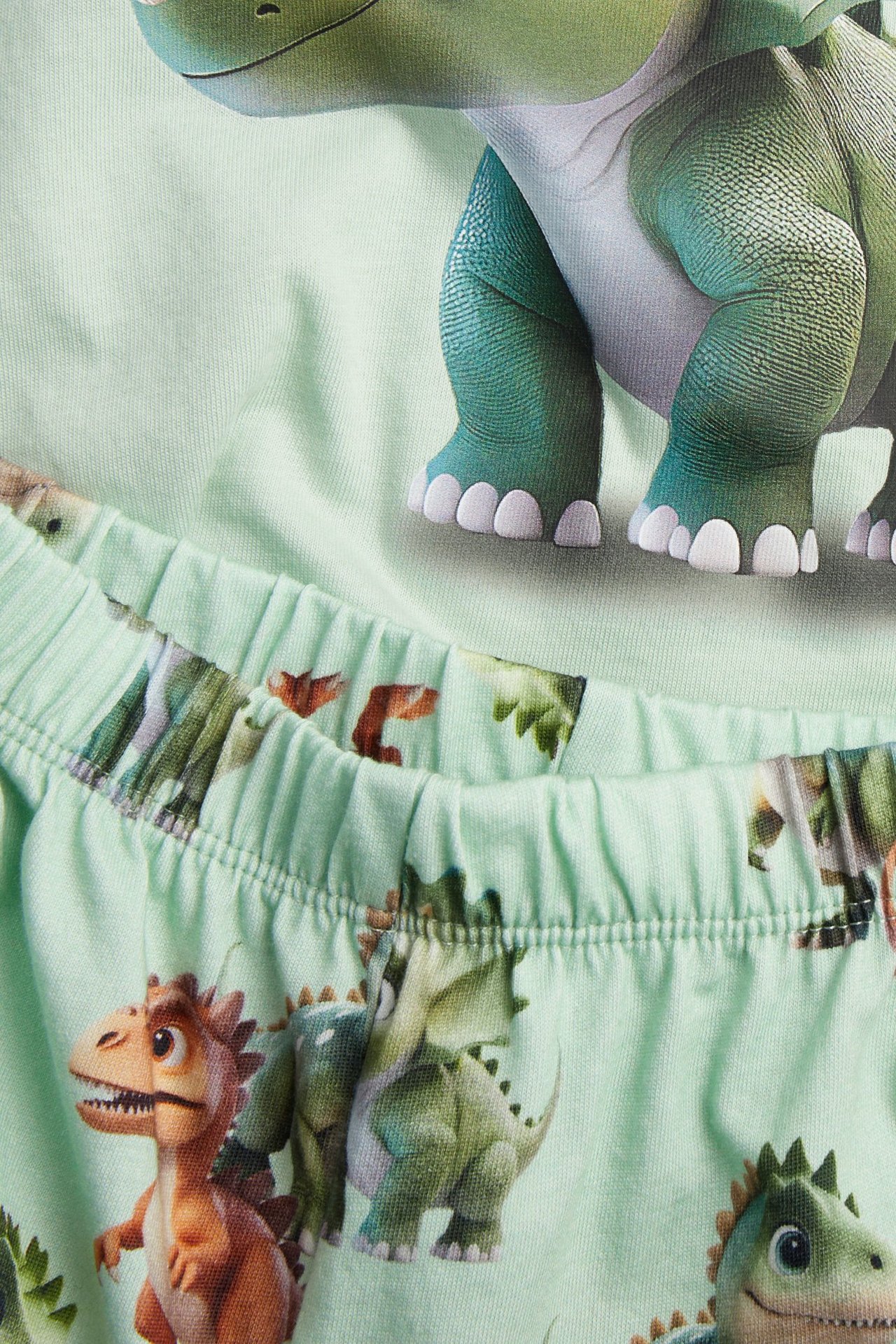 Närbild av ett grönt pyjamasset i bomull för barn med dinosauriemönster på tröjan och resårbyxorna.