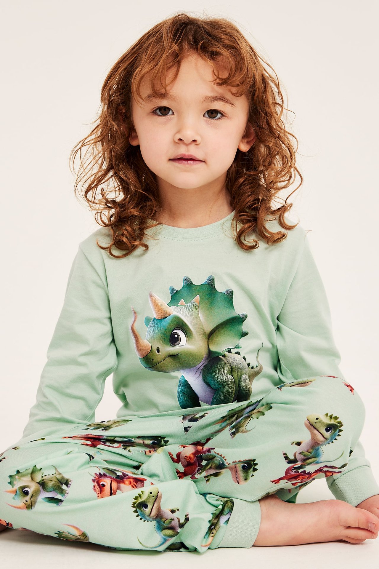 Edestä: Lapsi vaaleanvihreässä puuvillapyjamassa, jossa iso dinosaurusprintti paidassa ja dinosauruskuvioiset housut.