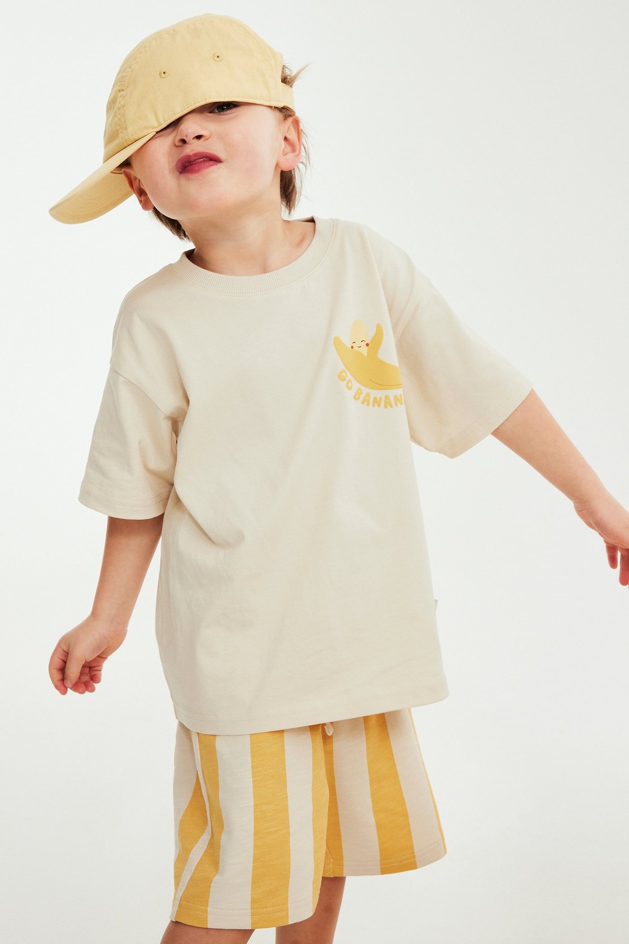 Edestäpäin: lapsi yllään beige t-paita banaanipainatuksella, keltaiset raidalliset shortsit ja keltainen lippalakki.