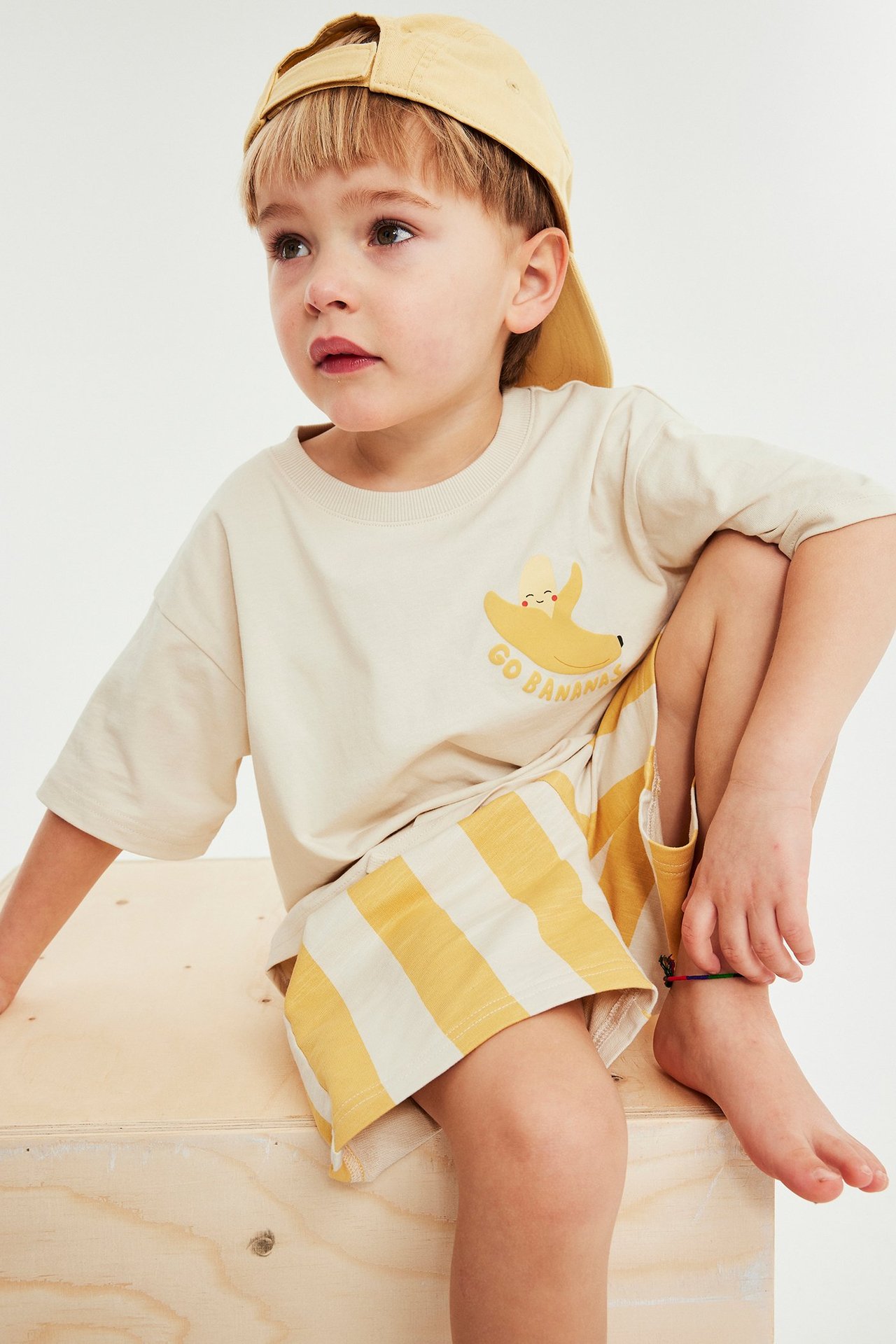 Framifrån: barn i beige t-shirt med banantryck, gula randiga shorts och gul keps.